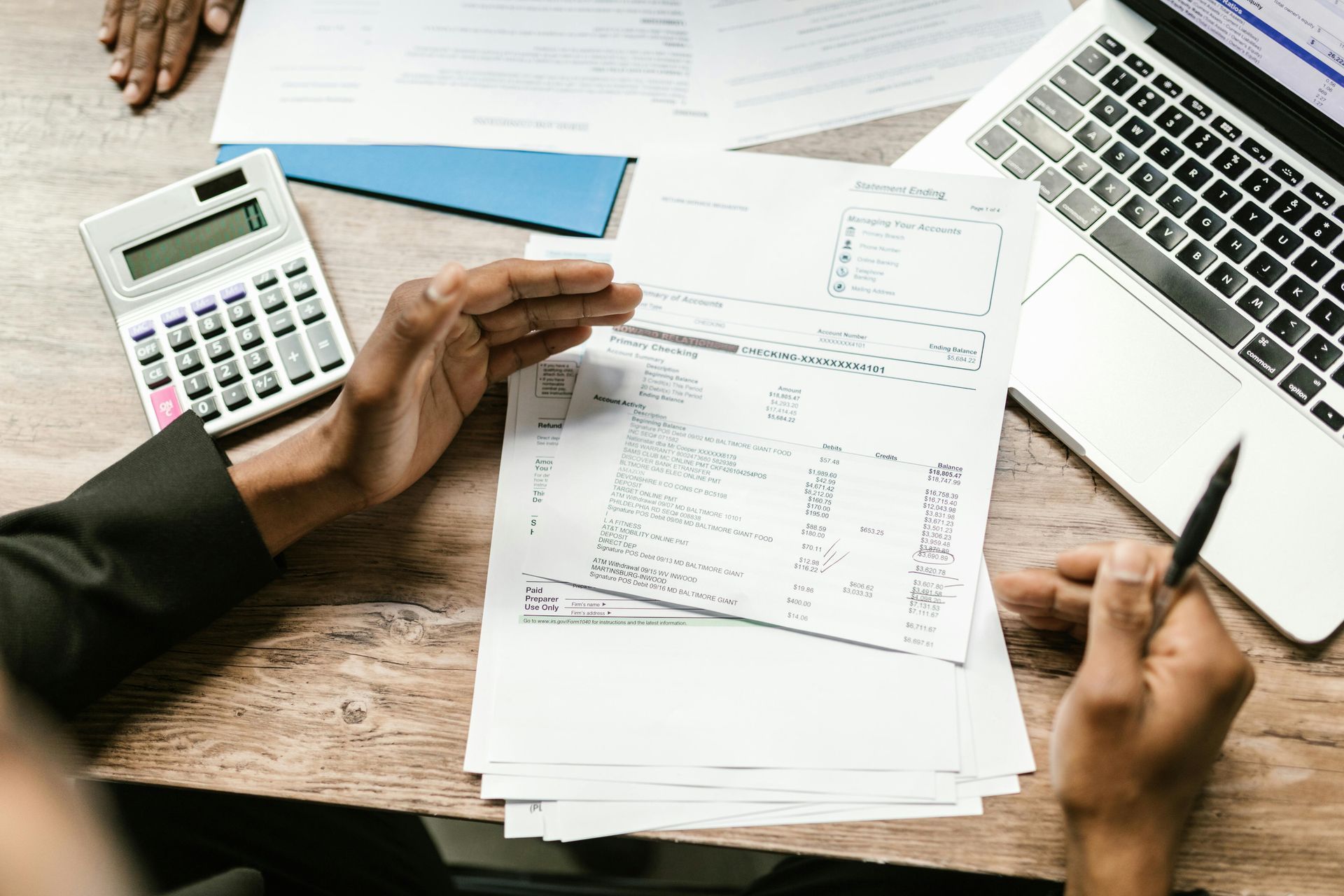 Person reviewing documents with calculator and laptop on a wooden desk.