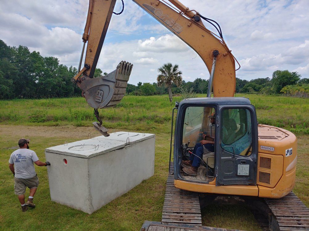 An excavator lifting a concrete septic tank with a worker guiding it in a grassy field.