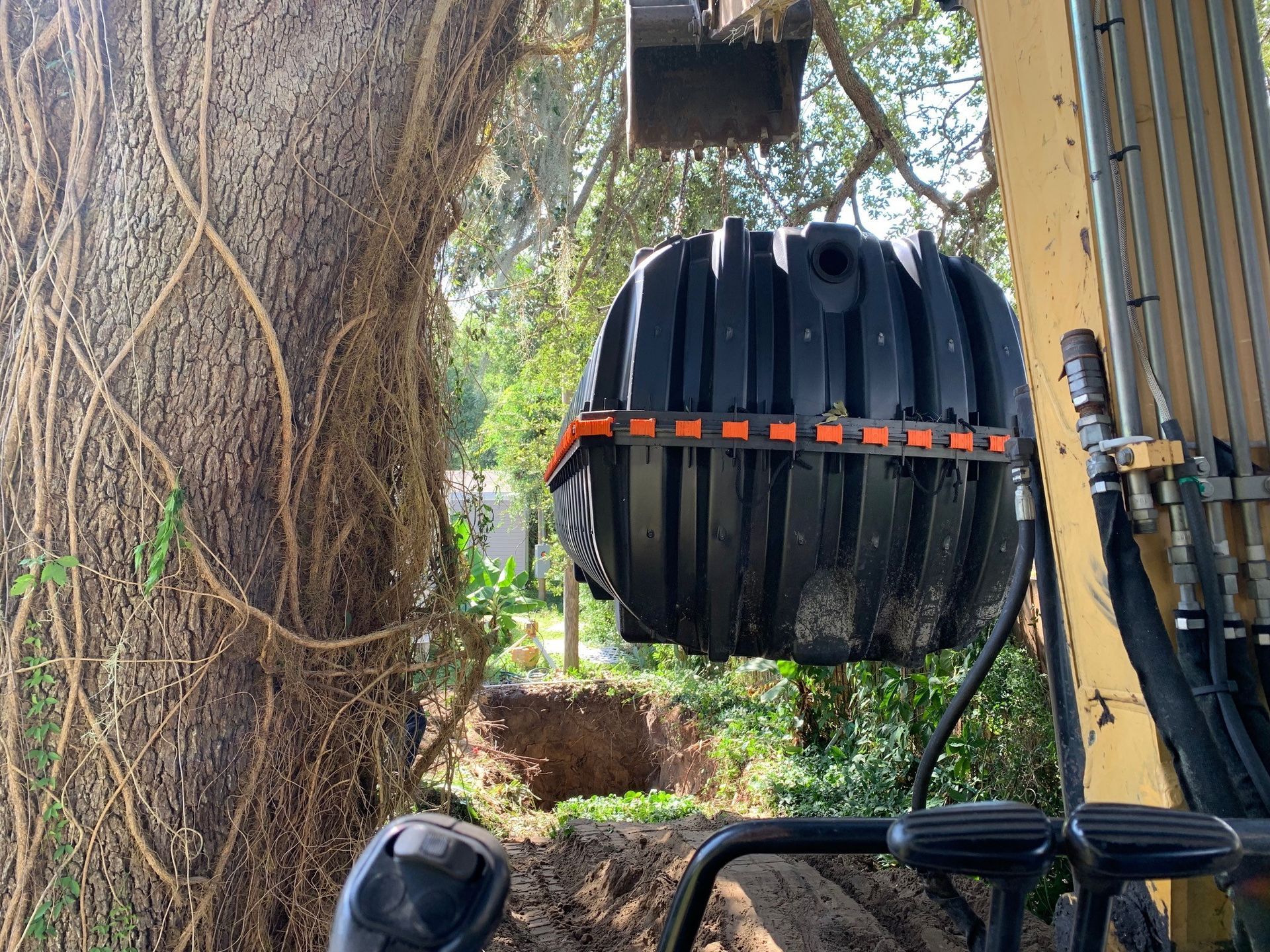 An excavator lifts a black, cylindrical tank near a tree. A hole is dug in the ground.
