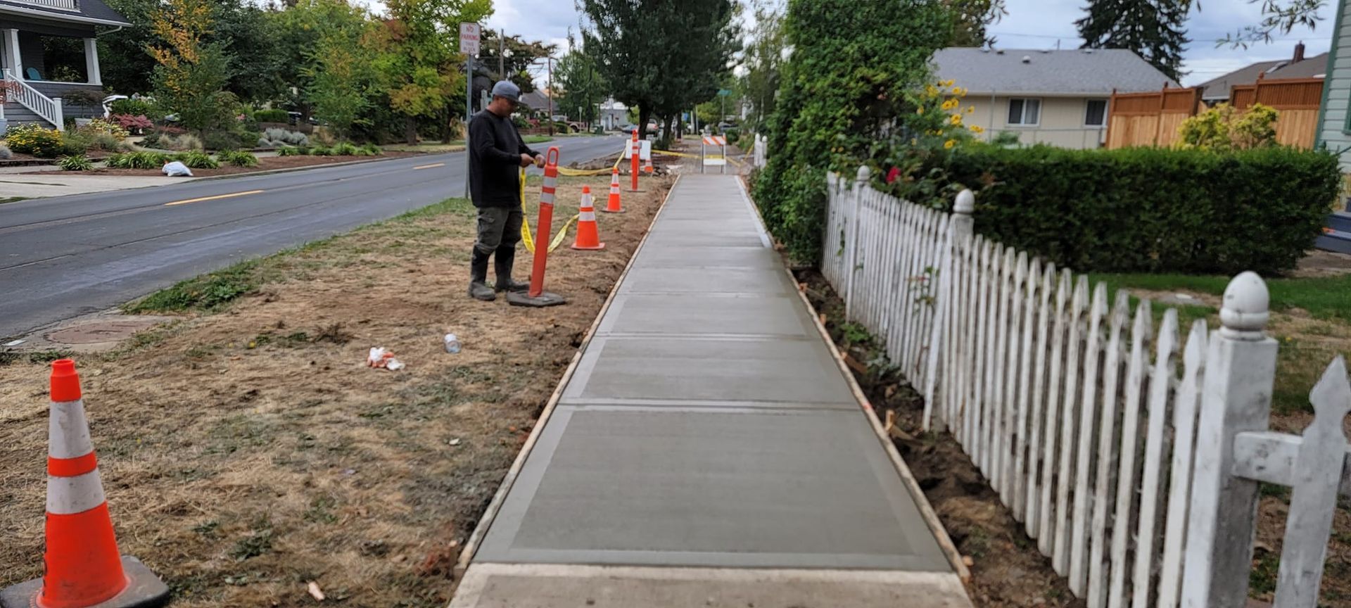 A man is standing on a sidewalk next to a white picket fence.