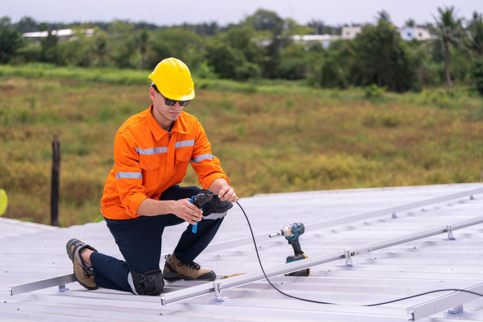 Person in safety gear installs solar panels on a rooftop, using a drill and kneeling.