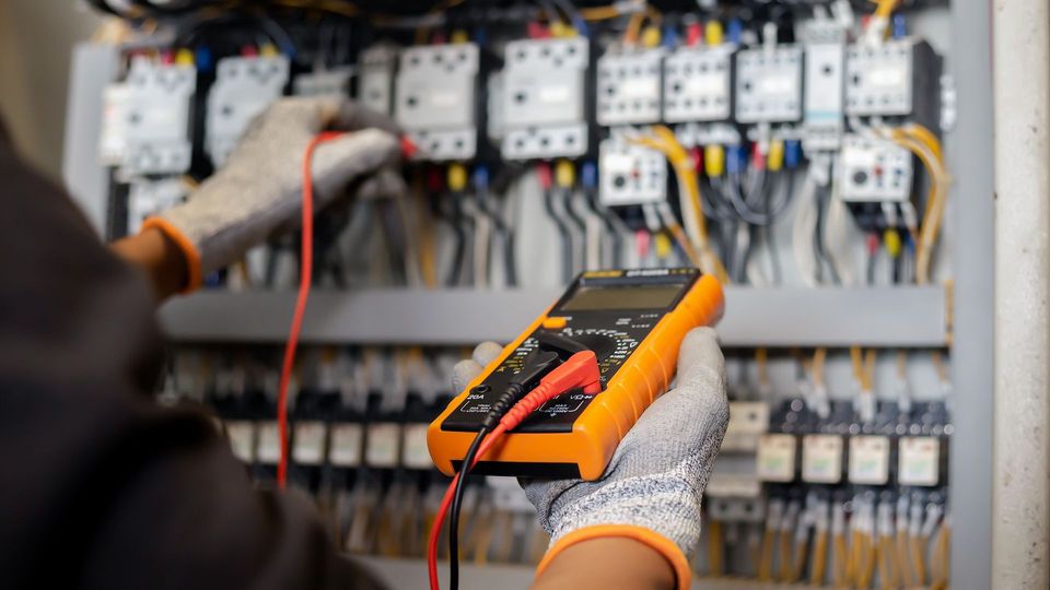 Electrician testing a control panel with a multimeter, wearing work gloves.