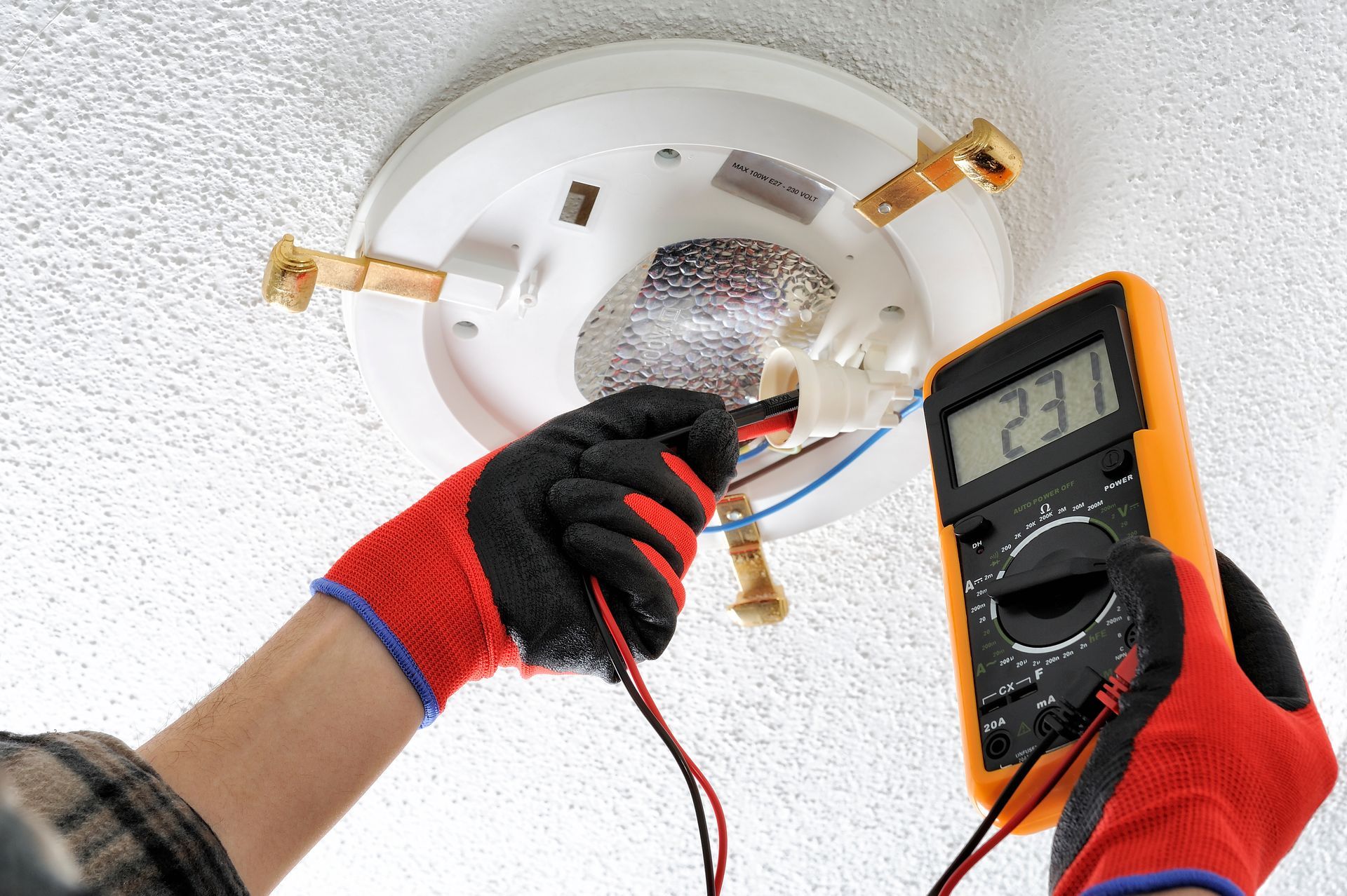 Person wearing red-and-black gloves tests wiring with a multimeter near a ceiling light fixture; digital display reads 23.1.