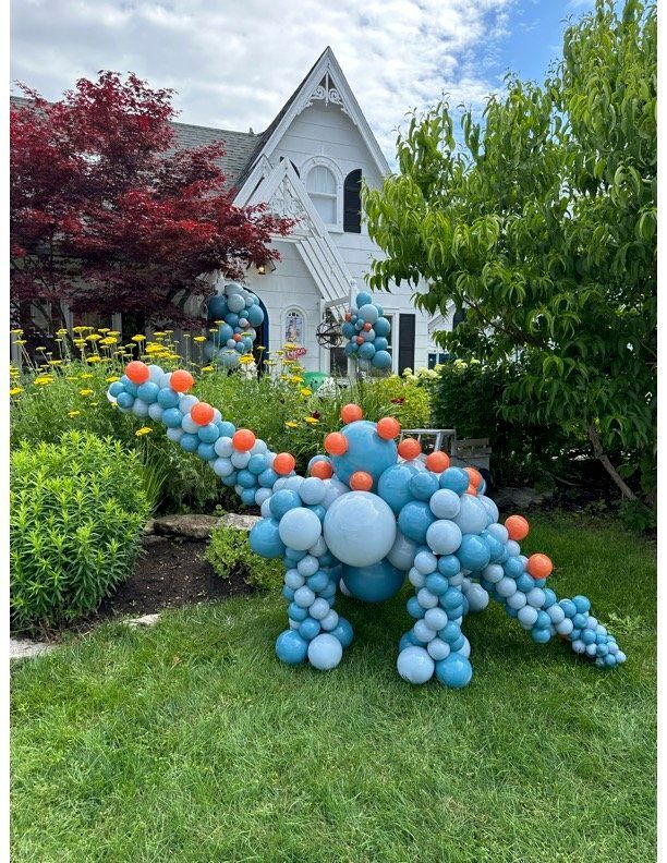 A dinosaur made out of balloons is sitting in the grass in front of a house.