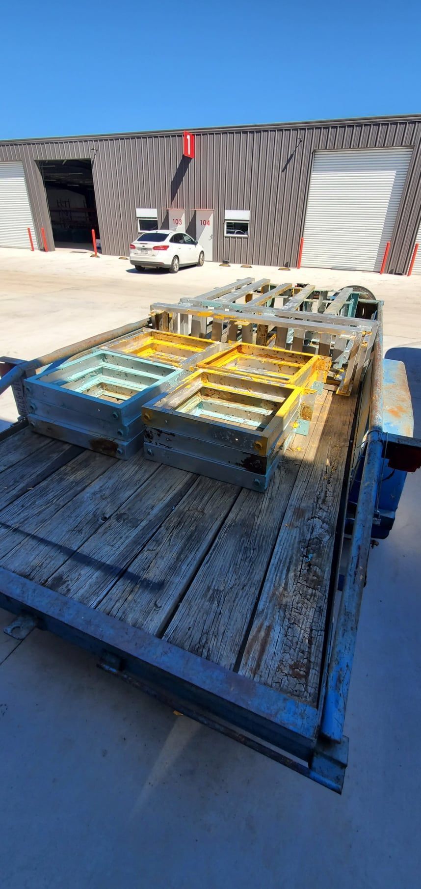 Wooden trailer carrying metal structures in front of a building with roll-up doors and a white car.