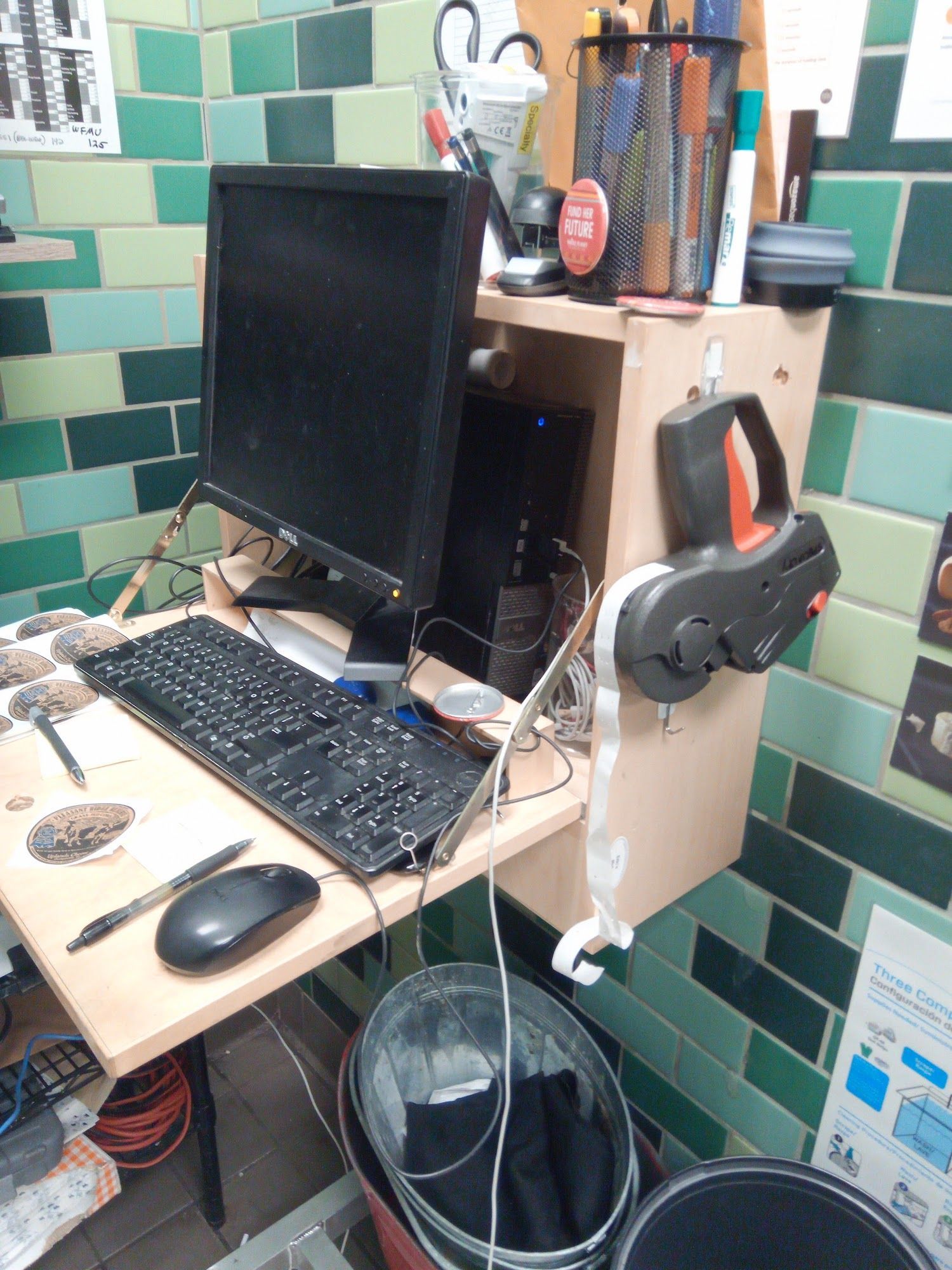 A cluttered computer setup: monitor, keyboard, mouse on a makeshift desk, against green tile.