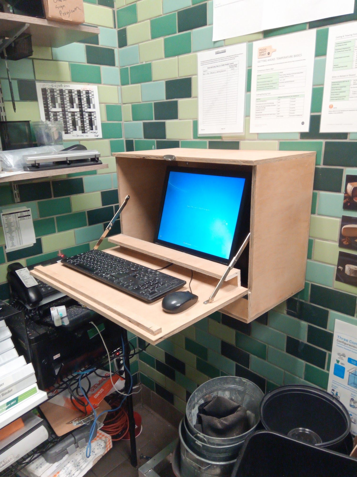 A computer with a keyboard and mouse mounted on a wall with a drop-down desk, surrounded by green tiles.
