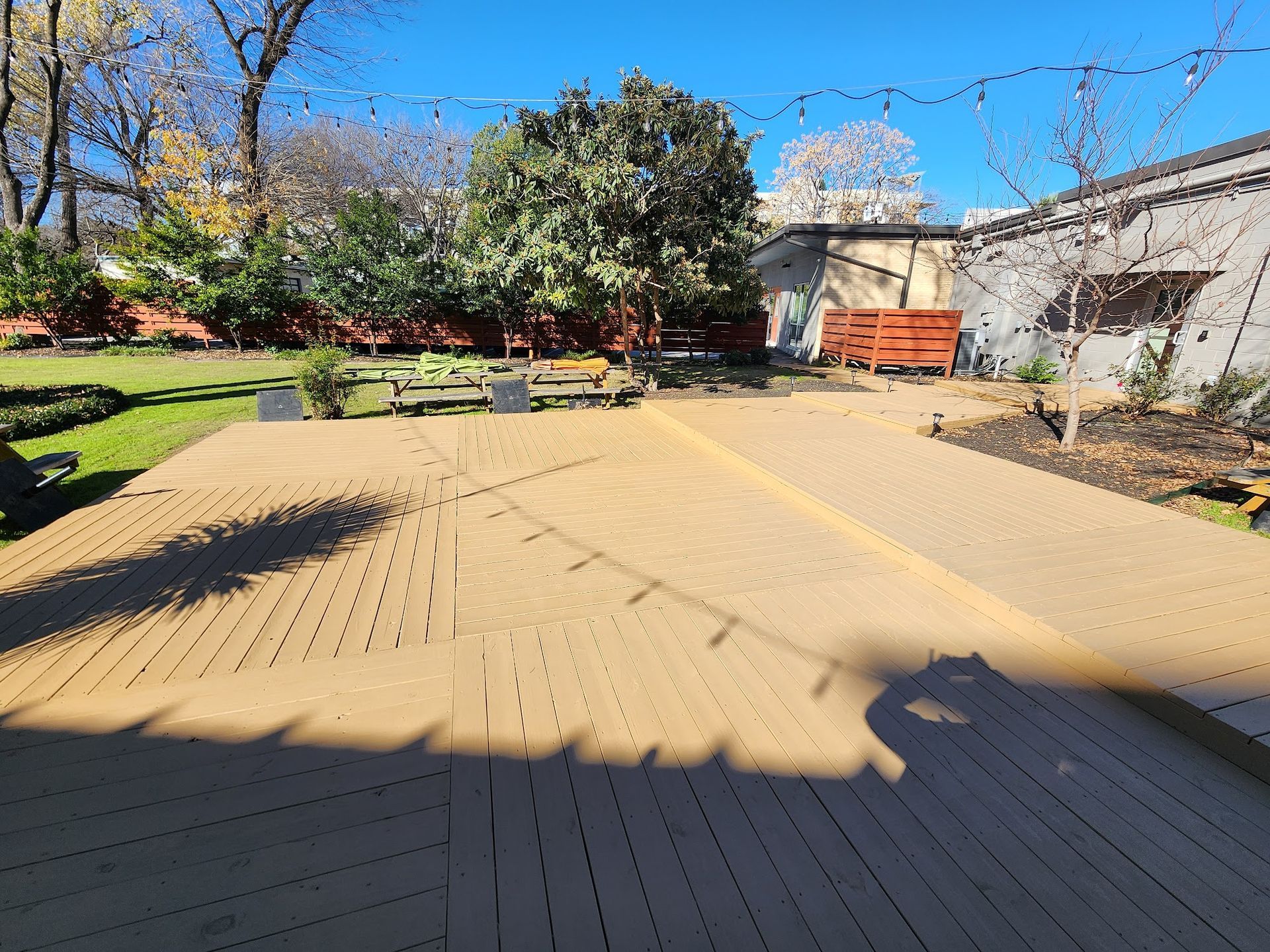 Wooden deck in a backyard with a tree and a small building under a clear sky.