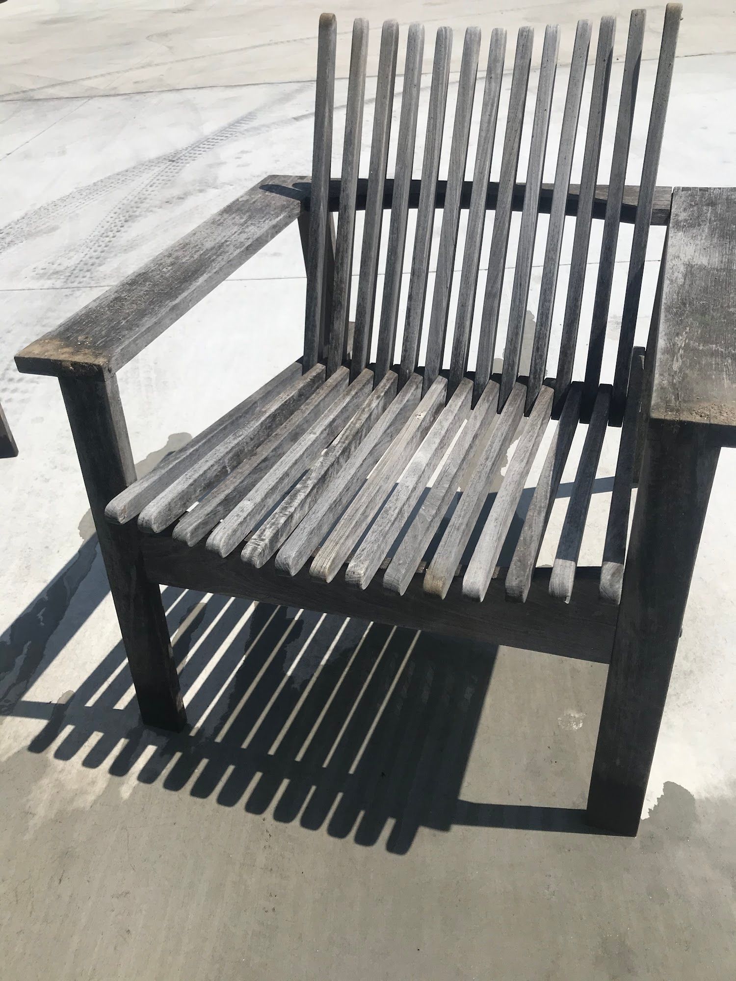 Wooden slatted chair outdoors, casting long shadow. Greyed wood, simple design.