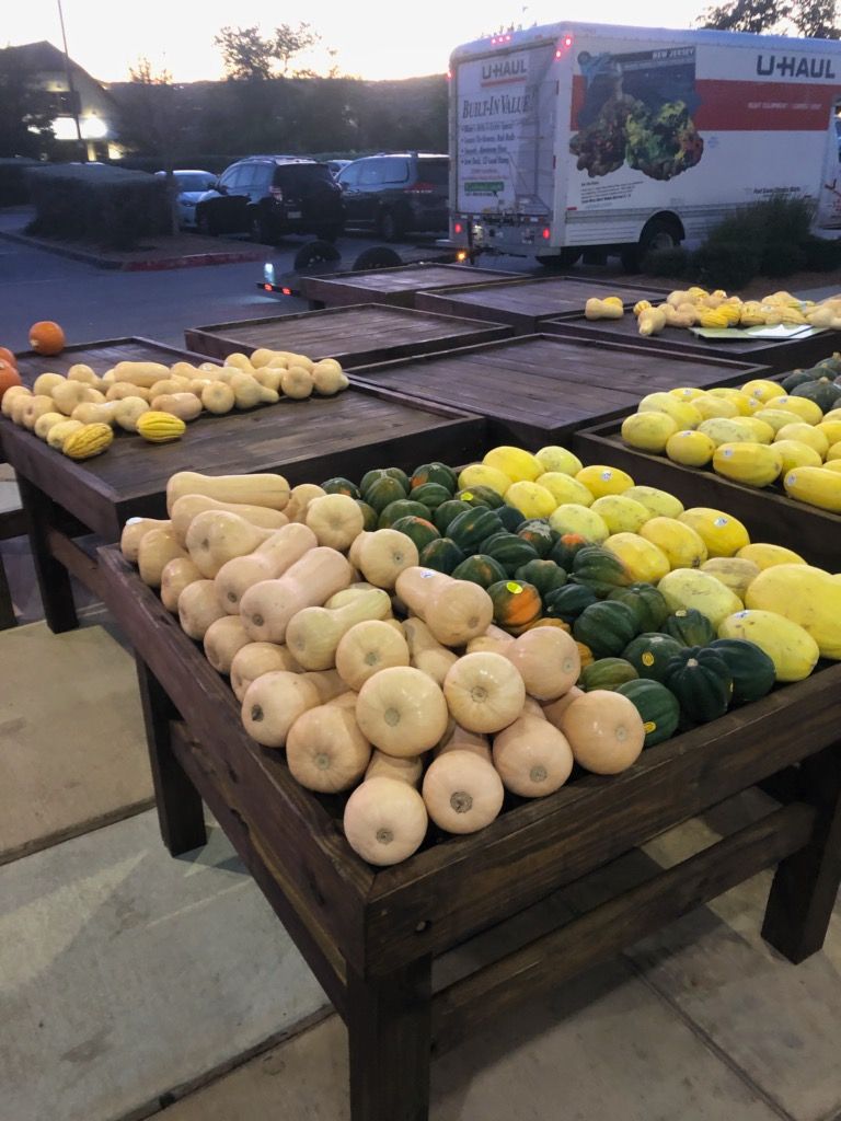 Wooden tables displaying various colorful squash at an outdoor market, with a U-Haul truck in the background.