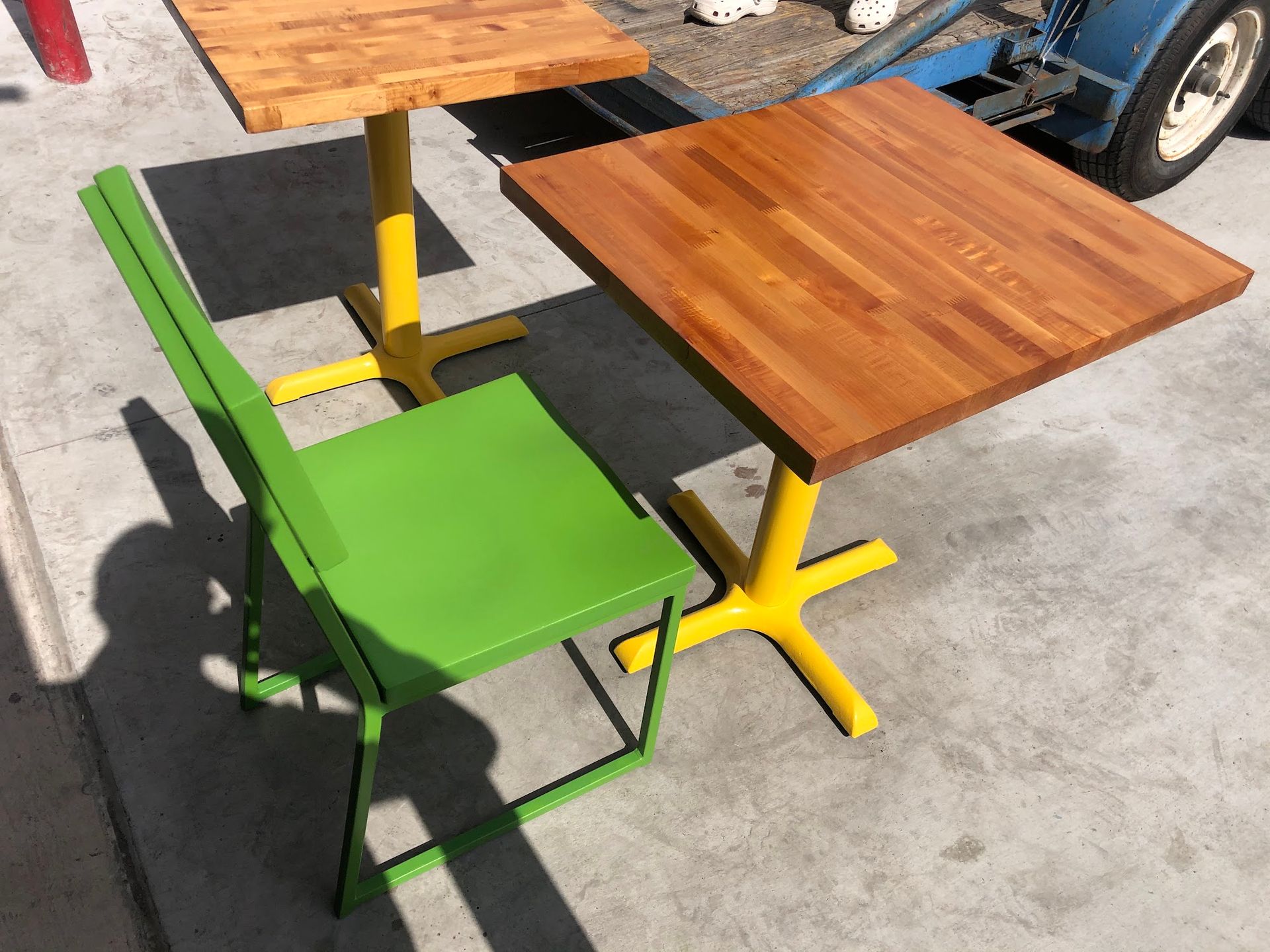 Green chair, two tables with yellow bases, and wooden tops; outdoors in sunlight.