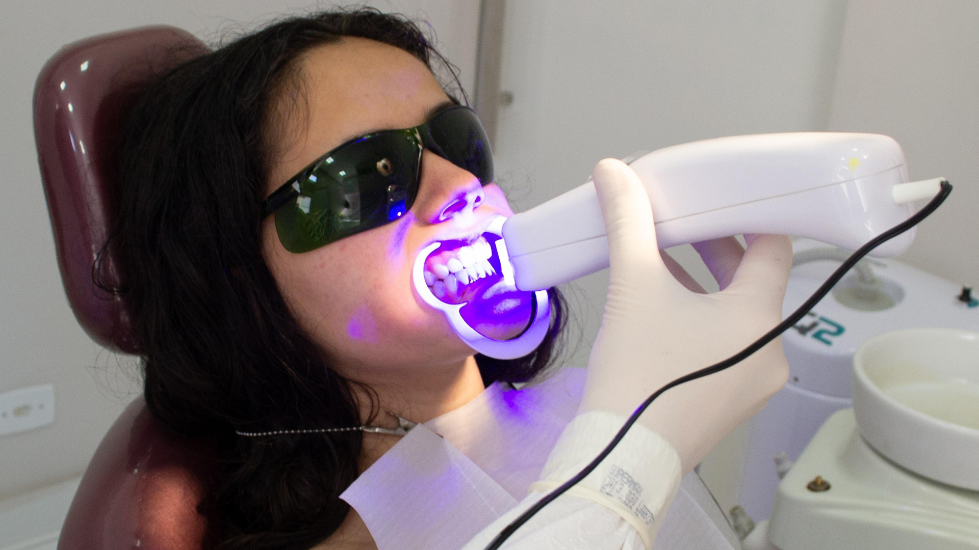Dentist using a curing light on a patient's teeth. Patient wears glasses, mouth open. Interior, dental office.