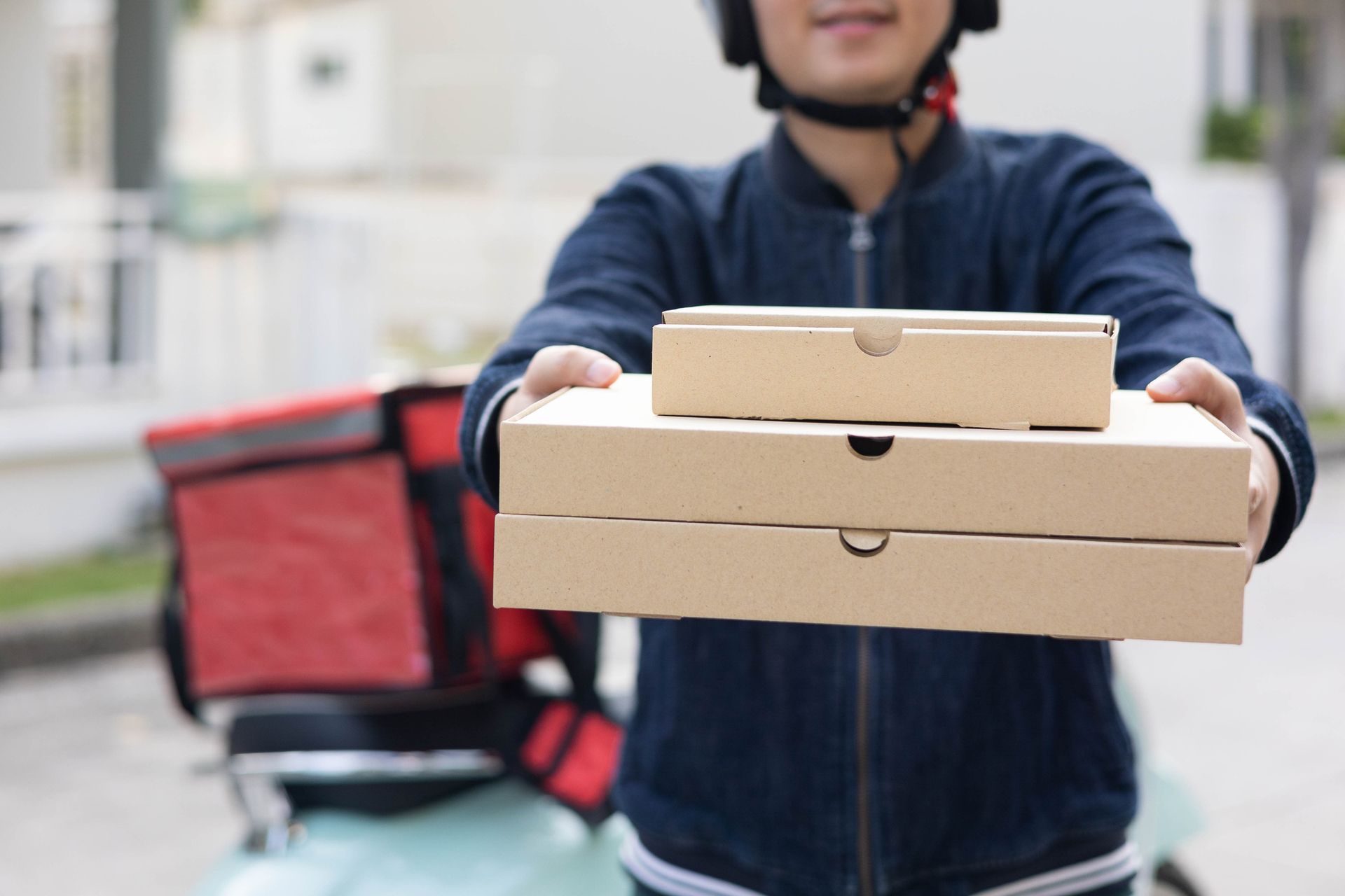 Delivery person holding pizza boxes with red delivery bag and scooter in urban background.