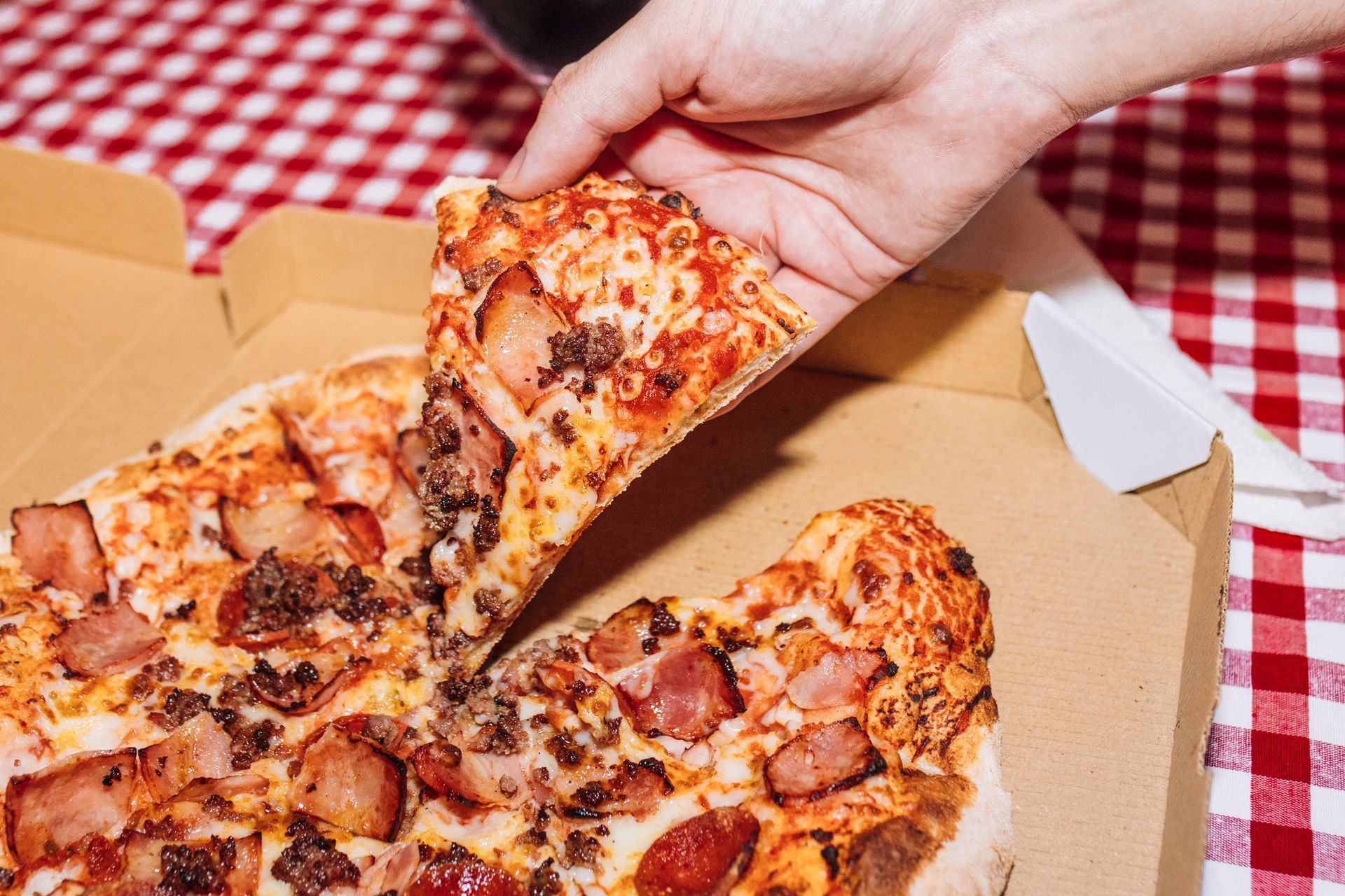 Hand picking up a slice of meat pizza from a cardboard box on a checkered tablecloth.
