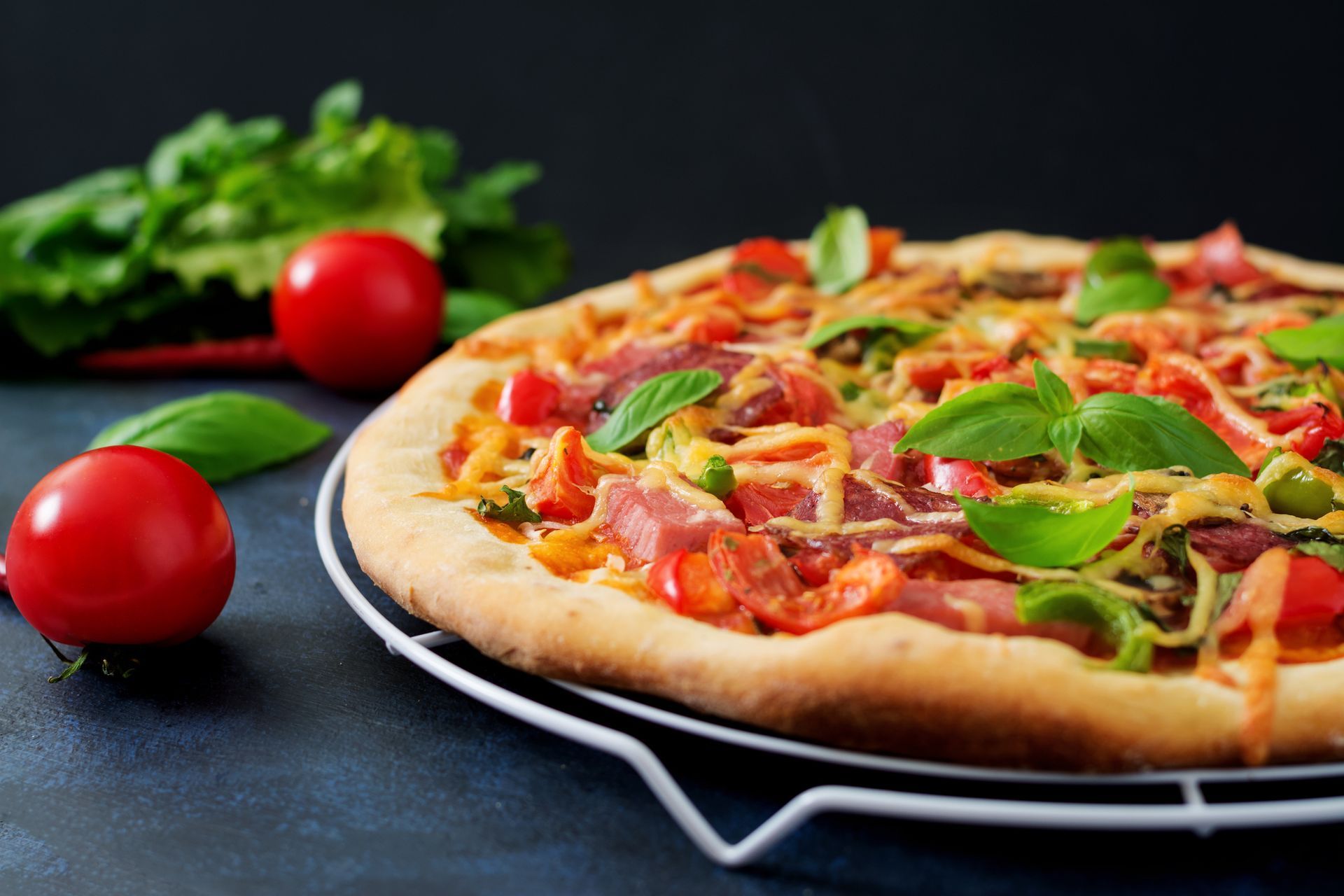 Pizza on a wire rack, garnished with fresh basil and tomatoes, with a dark background.