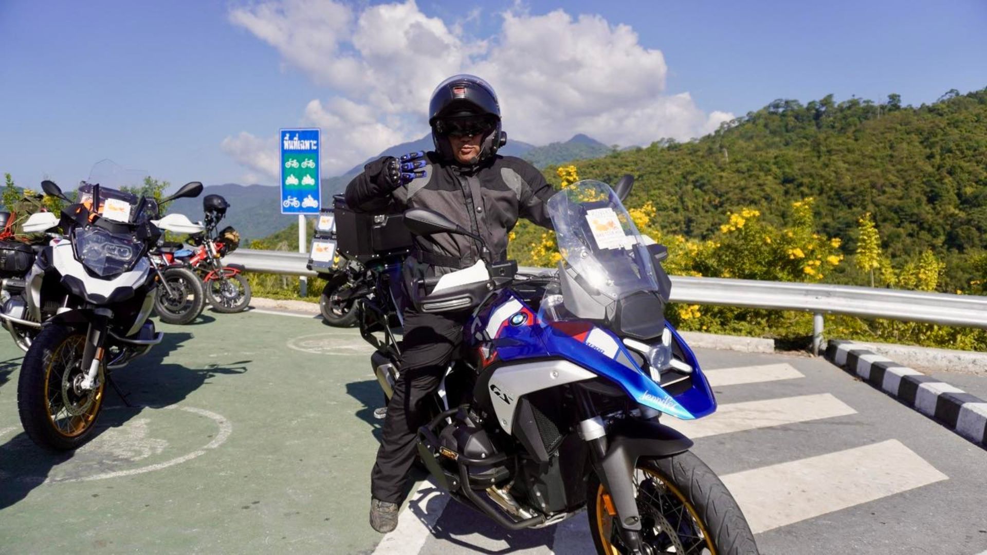 Man on a motorcycle giving a thumbs-up, mountain backdrop. Blue, white, and black motorcycle. Sunny day.