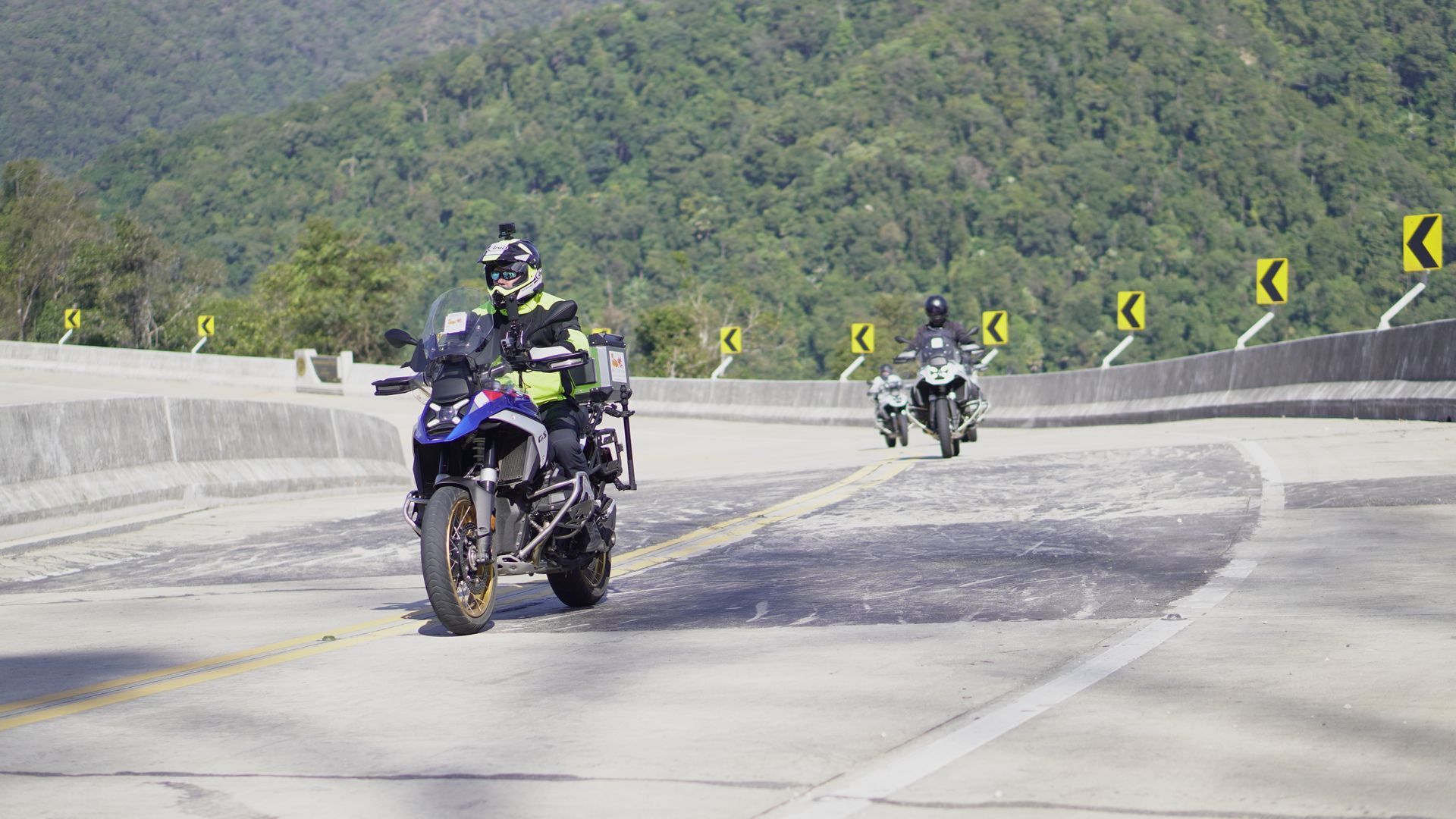Person sits on a motorcycle on a road near a green field and mountains. Motorcycle has luggage.
