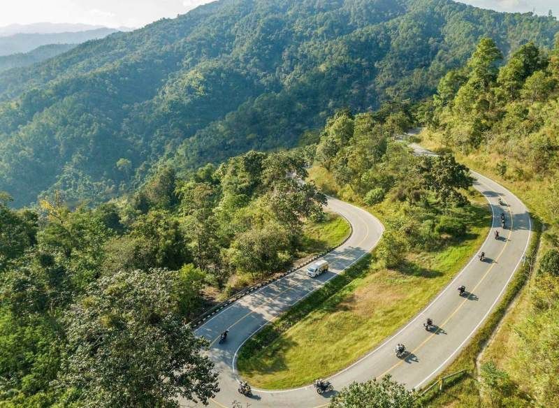 A drone view of motorcycle riders navigating sweeping mountain switchbacks surrounded by dense forest in northern Thailand