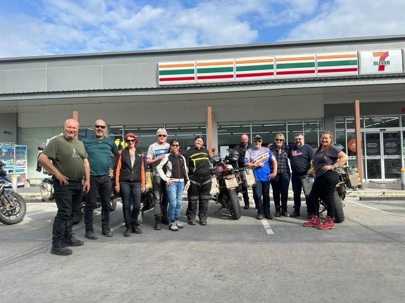 A group of motorcycle tour participants standing in front of their bikes outside a 7-Eleven store in Thailand, smiling and posing for a group photo