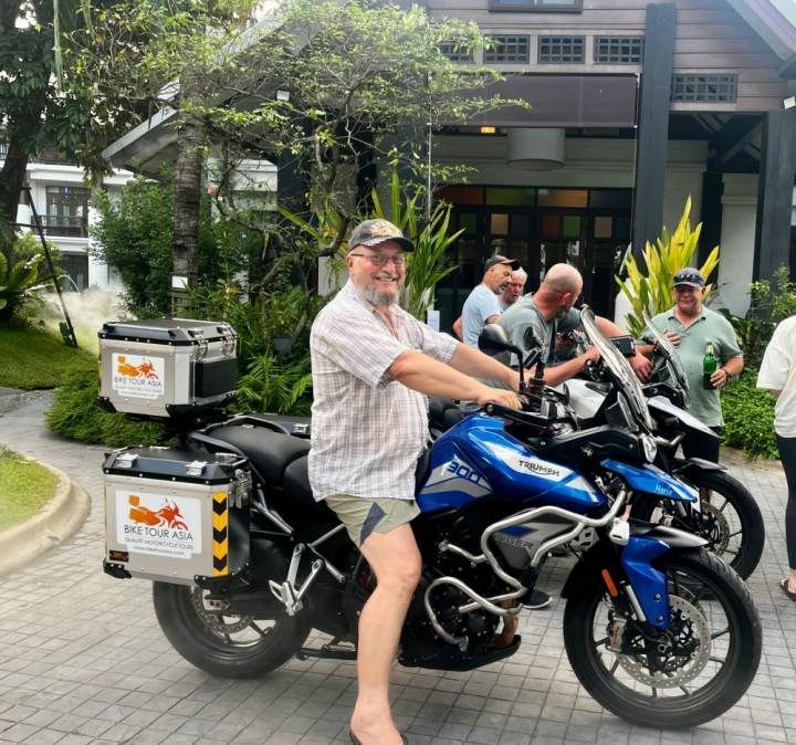 A smiling rider sitting on a Triumph Tiger motorcycle during a Bike Tour Asia trip in Thailand, with other riders gathered in the background.