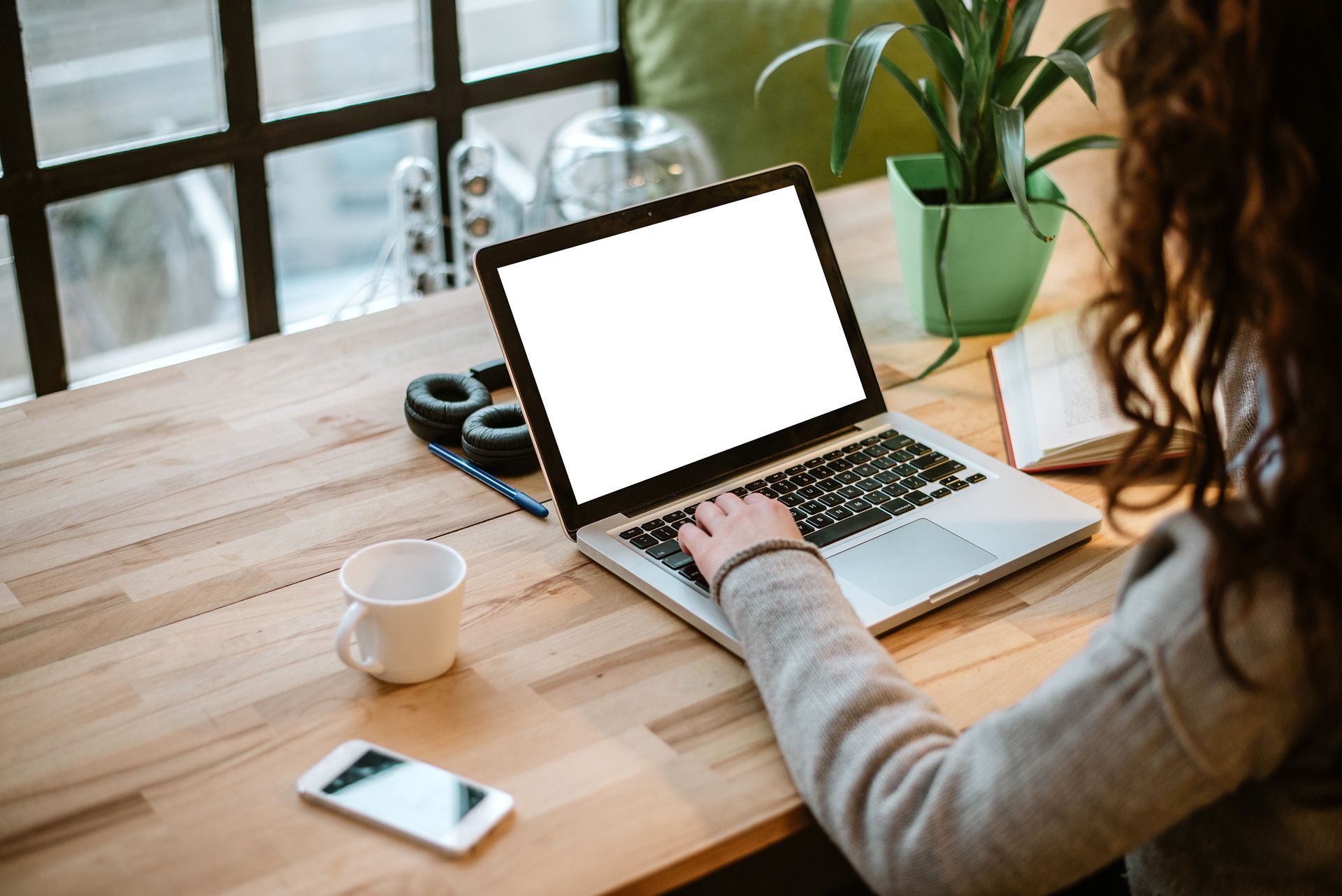 A person types on a laptop at a wooden desk near a window, with a plant, coffee mug, and phone nearby.
