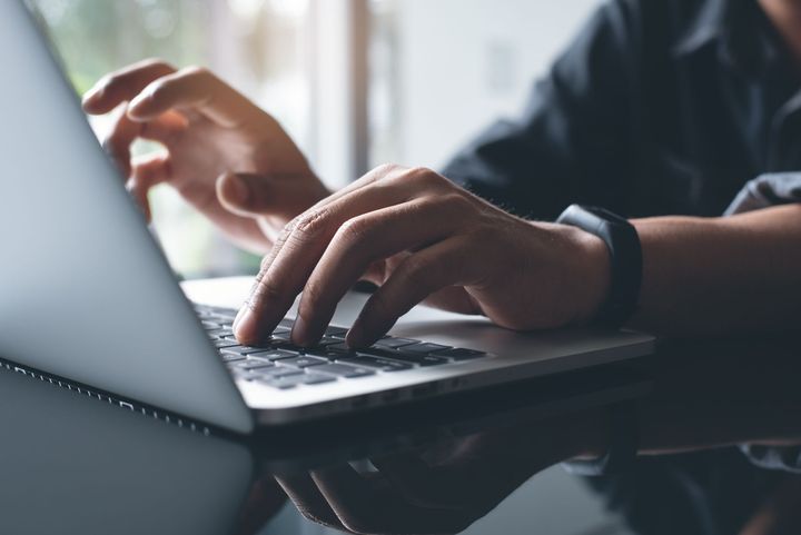 A person typing on a laptop computer, seen from a side angle, with a black fitness tracker visible on their wrist.