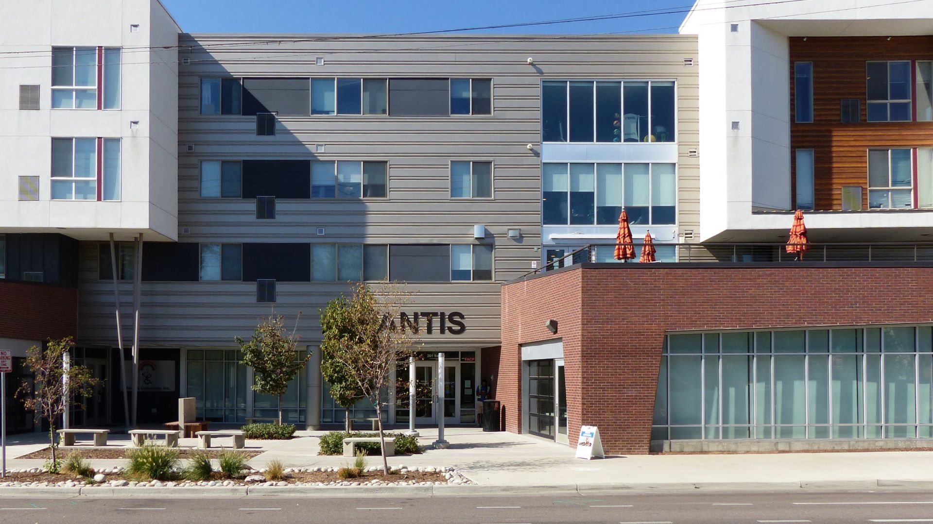 A modern four-story building with a brick-faced entrance, horizontal siding, and white paneling under a clear blue sky.