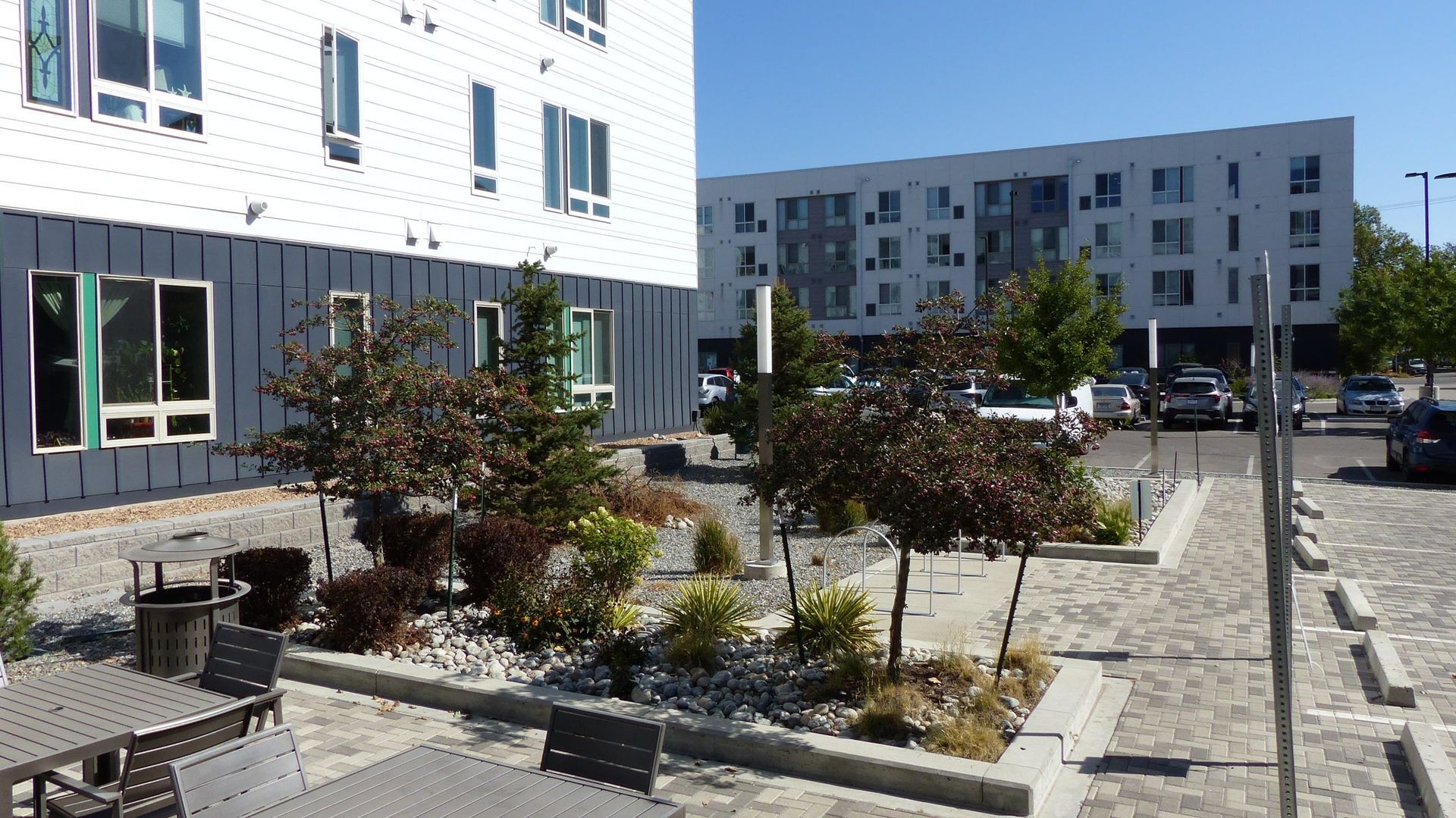 Outdoor patio seating with landscaping in front of a multi-story apartment building under a clear blue sky.