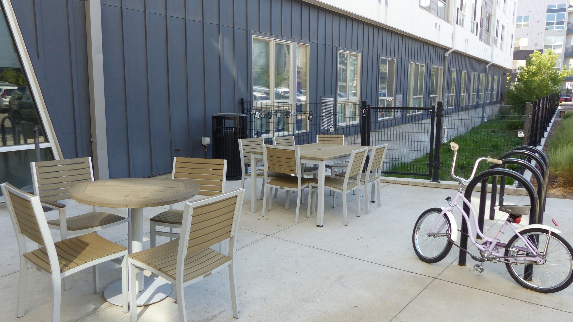 An outdoor patio with metal tables and chairs next to a bike rack with a light-colored bicycle in front of a blue building.