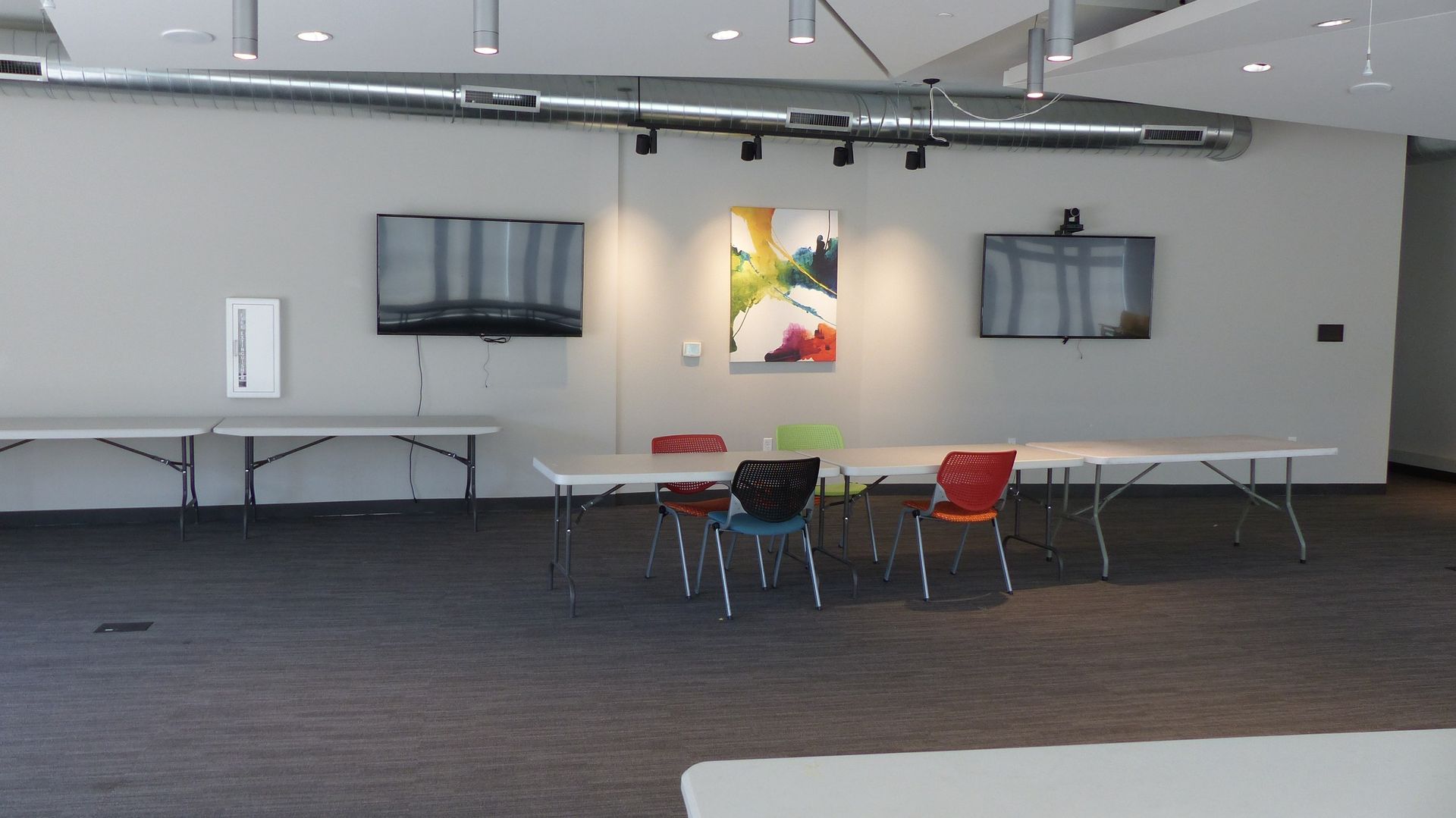 A meeting room with a long table, colorful chairs, two wall-mounted televisions, and abstract art on a white wall.