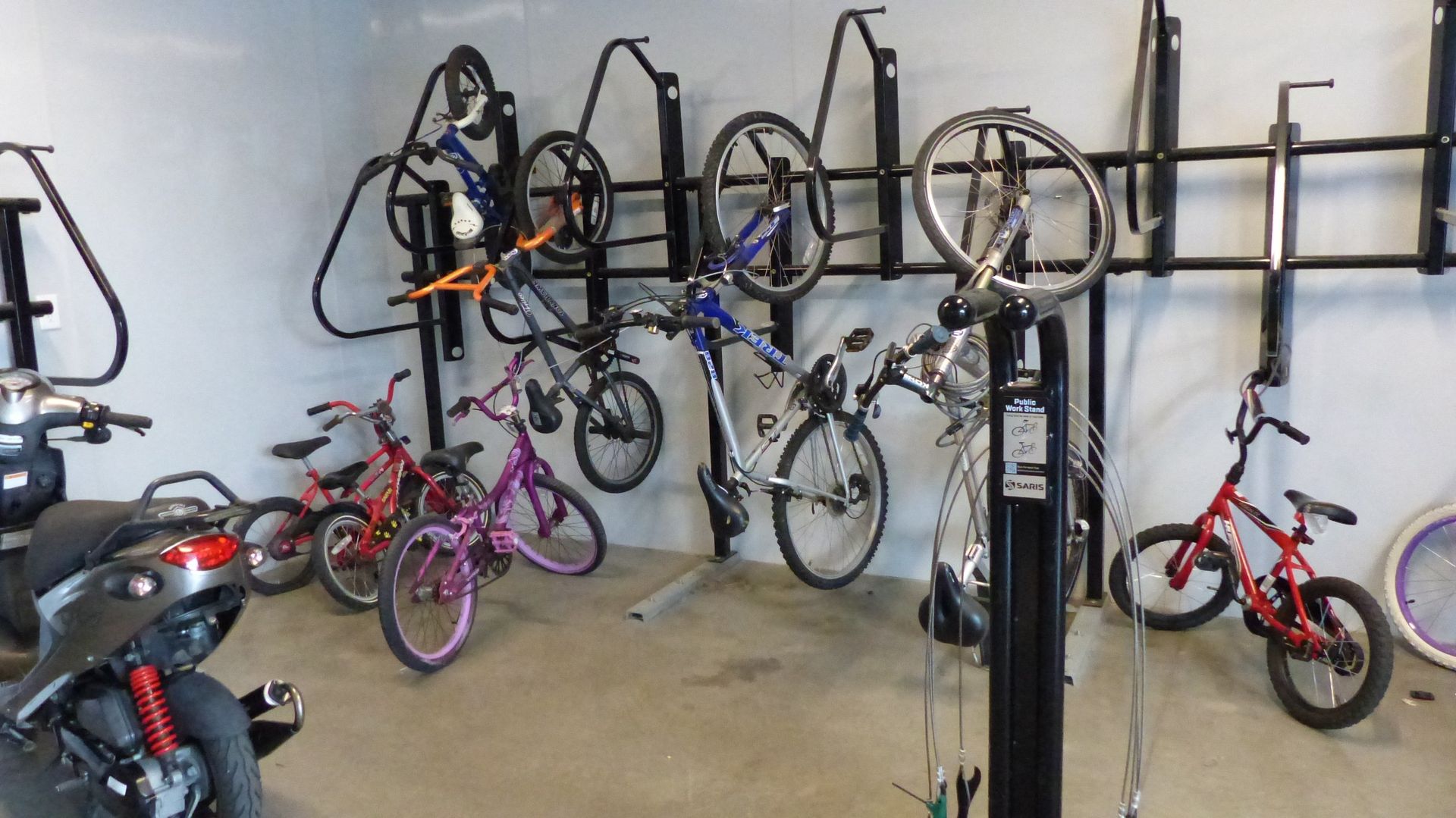 A storage area with several bicycles parked on the floor and mounted on a wall rack, plus a scooter in the foreground.