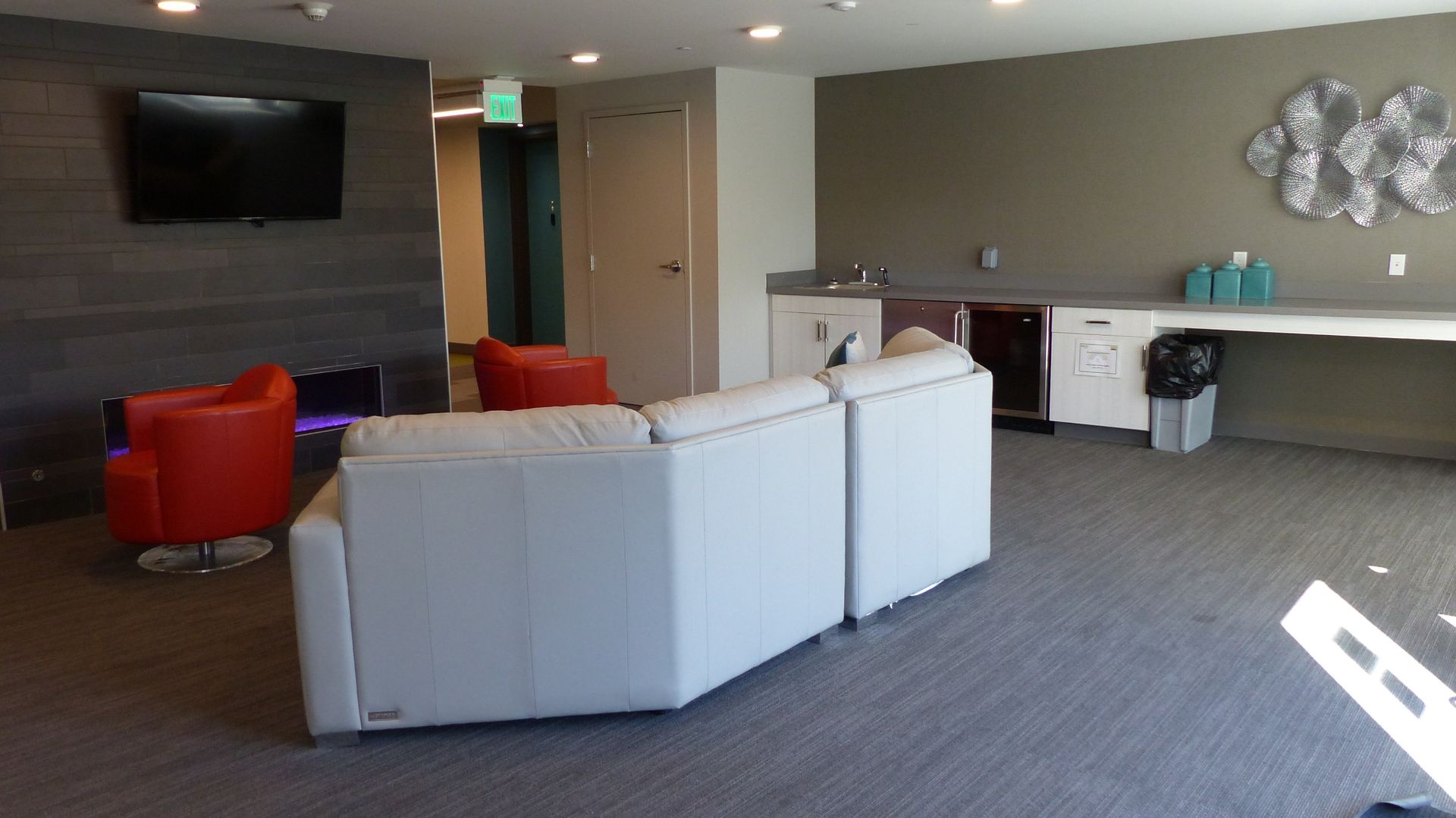 A modern lounge area with a white sectional sofa, orange armchairs, a wall-mounted TV, and a beverage station.