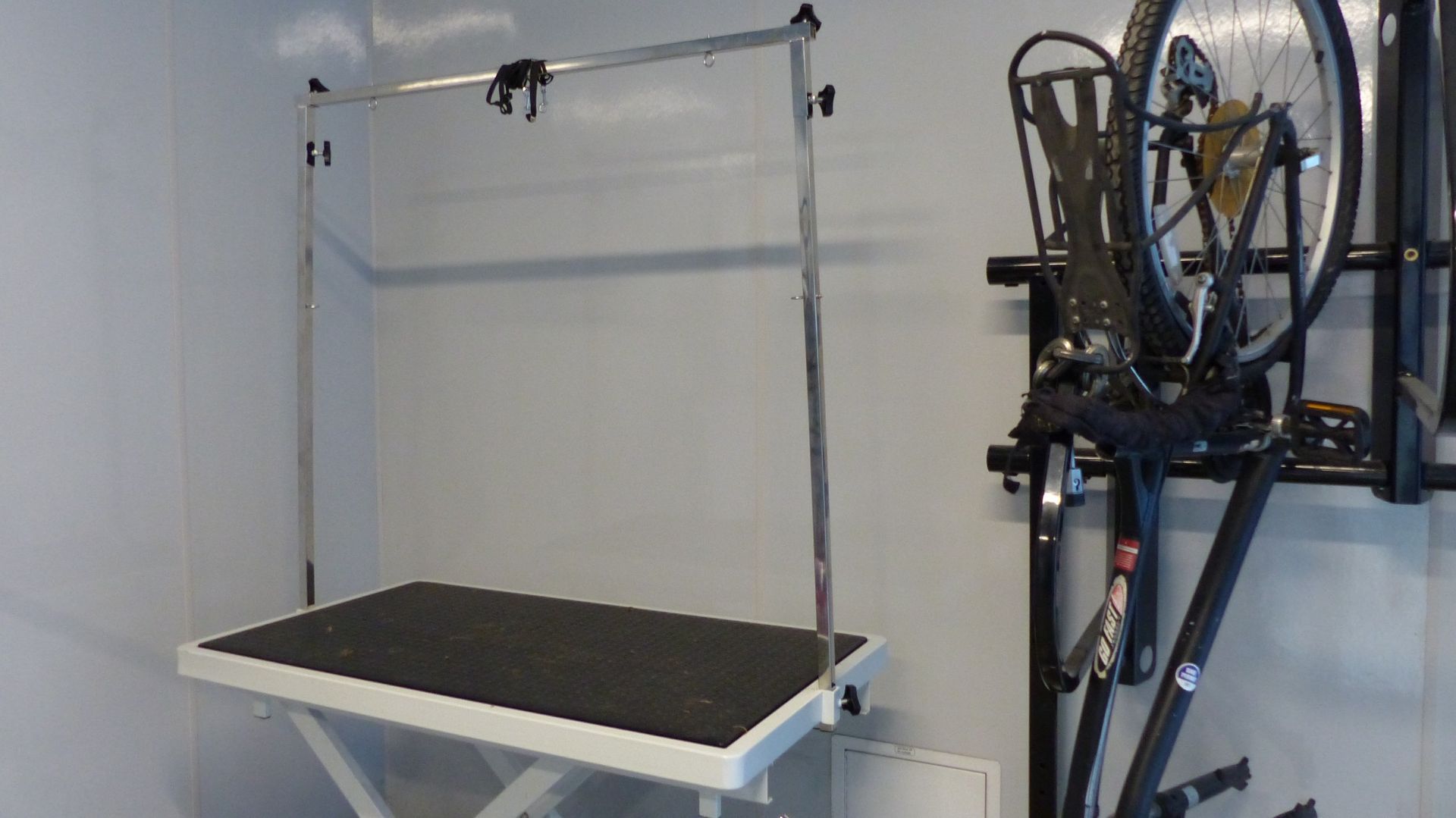 A grooming table with an overhead arm stands beside a black bicycle mounted on a wall rack in a garage or room.