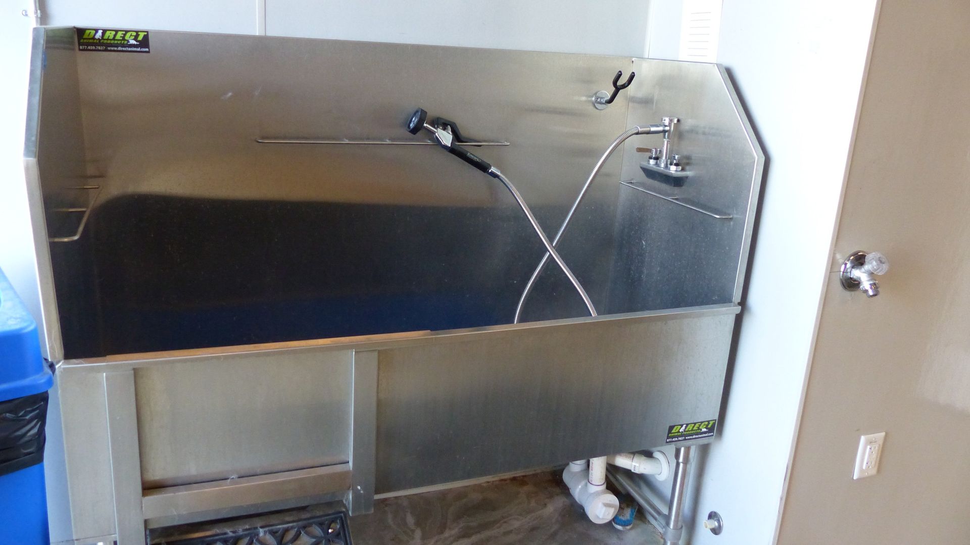 A stainless steel pet grooming tub with a spray nozzle and faucet, positioned against a white wall next to a blue bin.