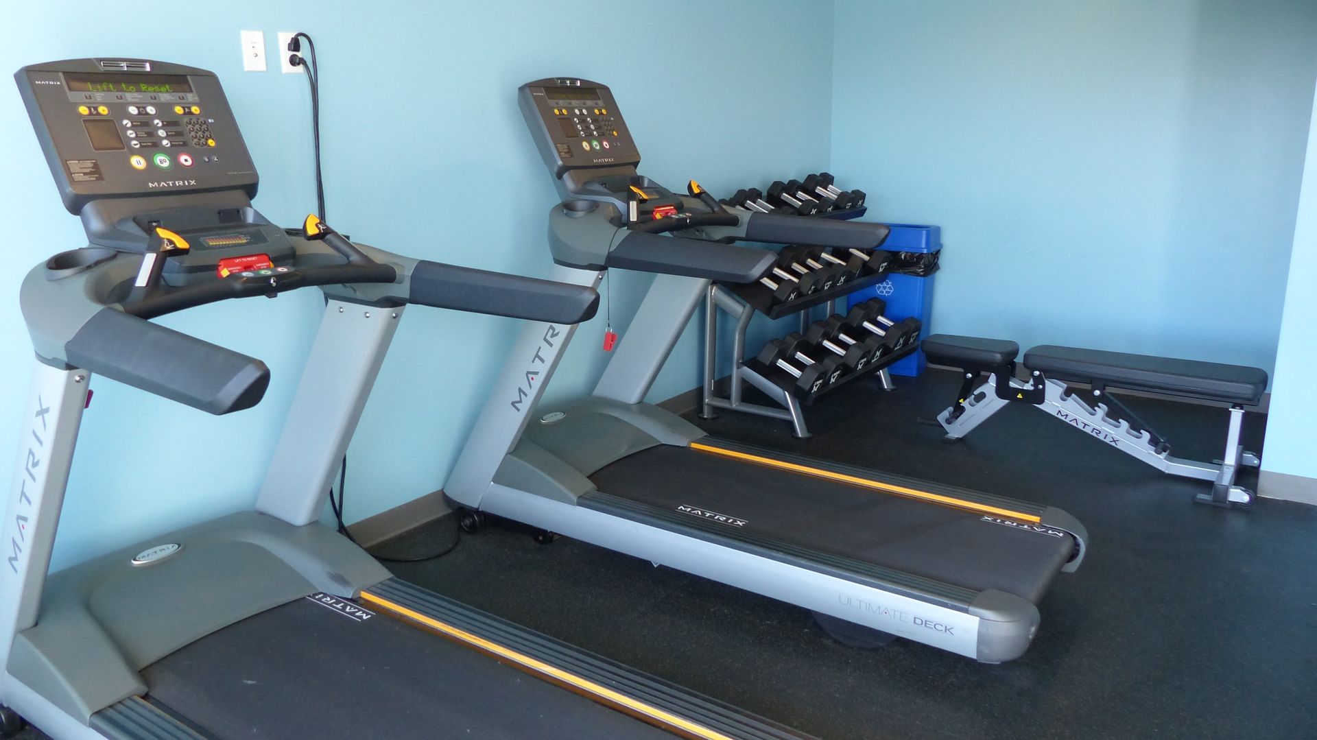 Two Matrix treadmills, a rack of dumbbells, and a weight bench in a gym with light blue walls.
