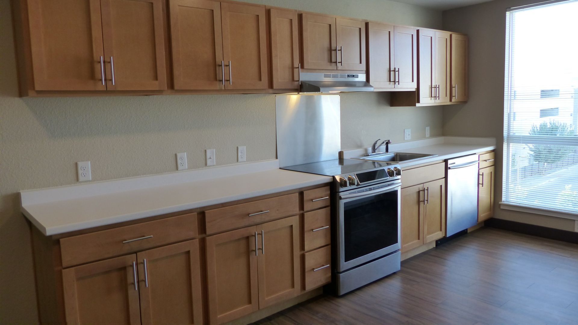 A modern kitchen with light wood cabinets, white countertops, stainless steel appliances, and a window to the right.