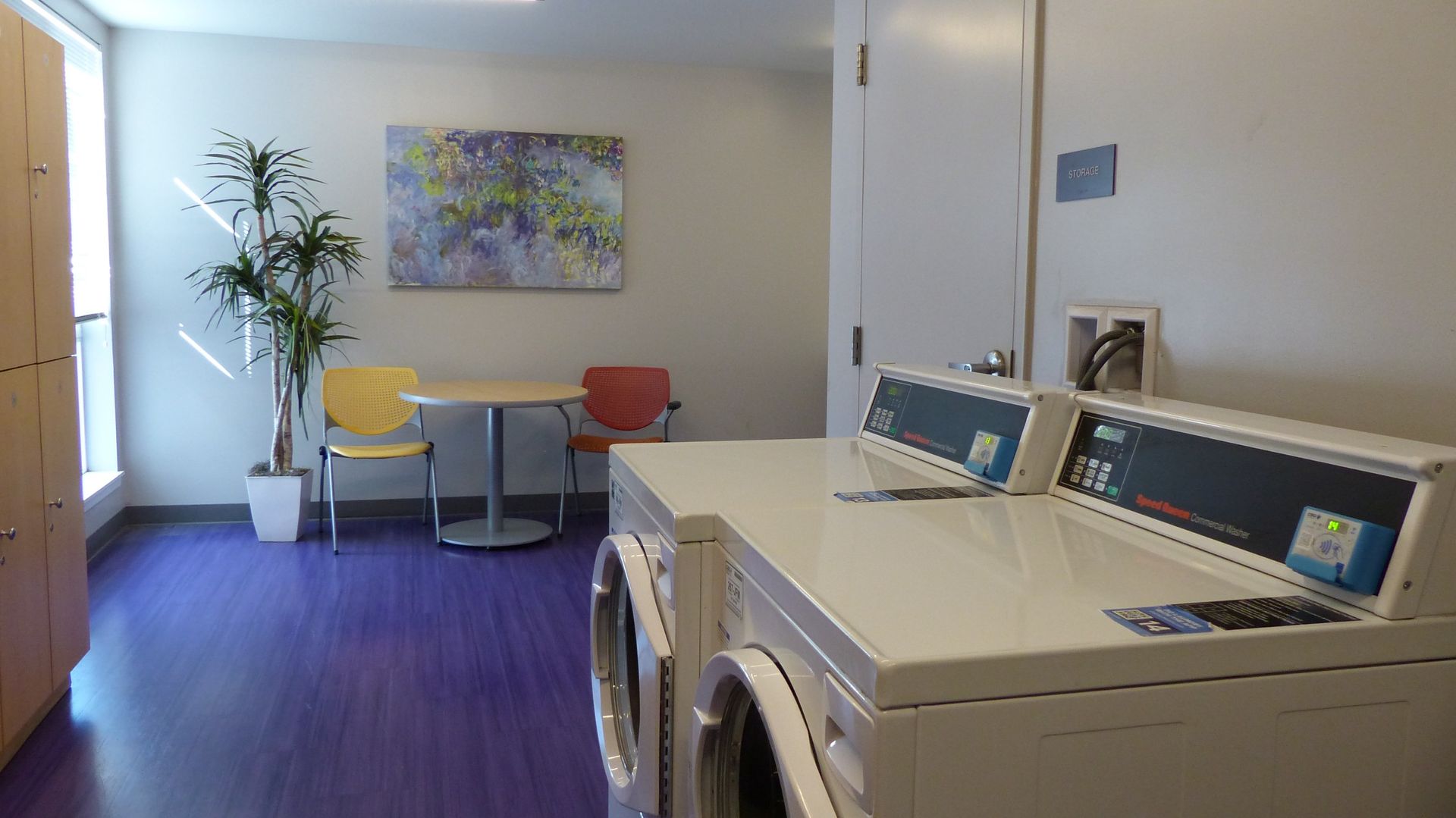 A laundry room with two white machines in the foreground, and a small table with yellow and orange chairs in the back.