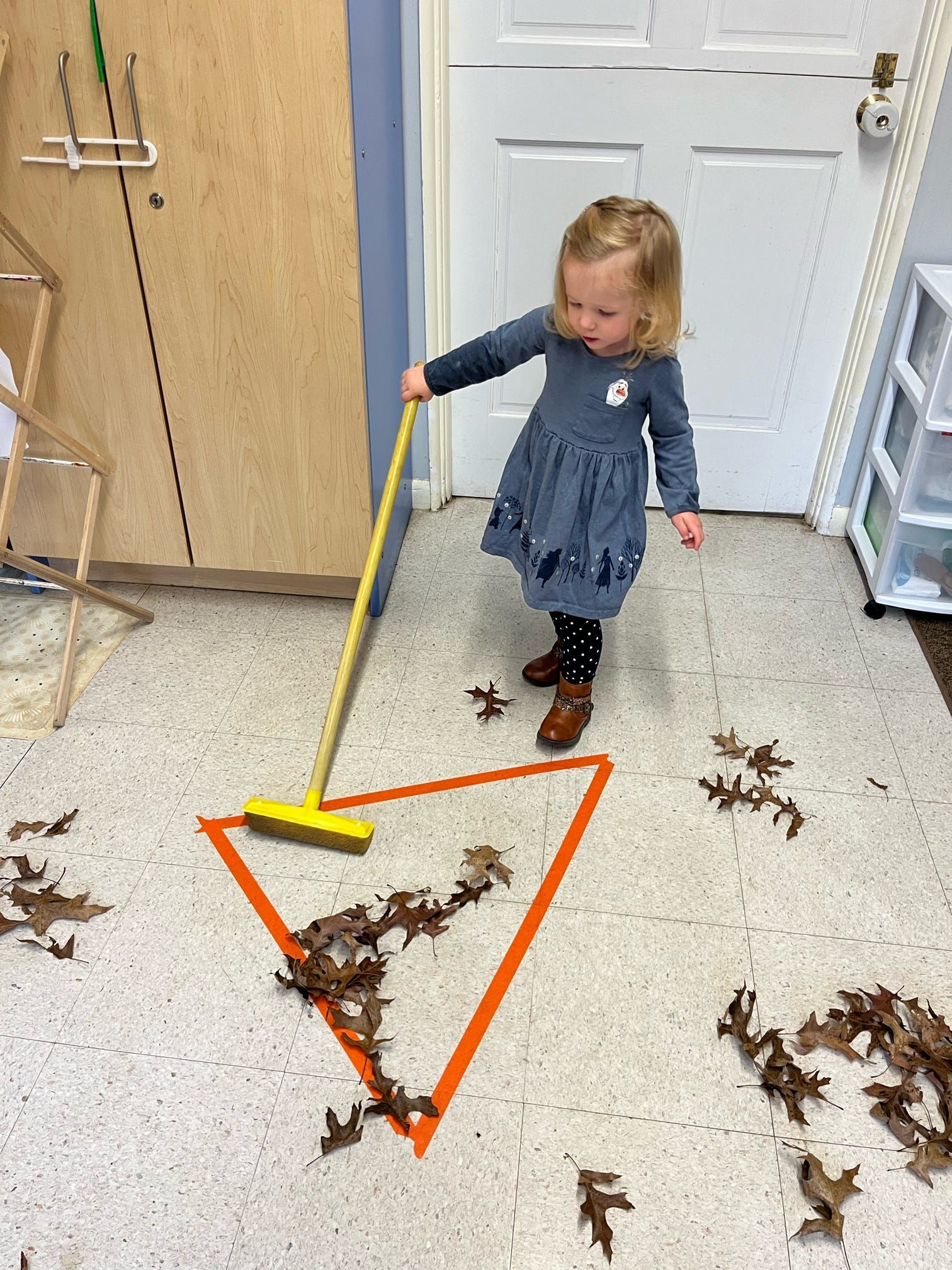 A little girl is sweeping leaves on the floor with a broom.