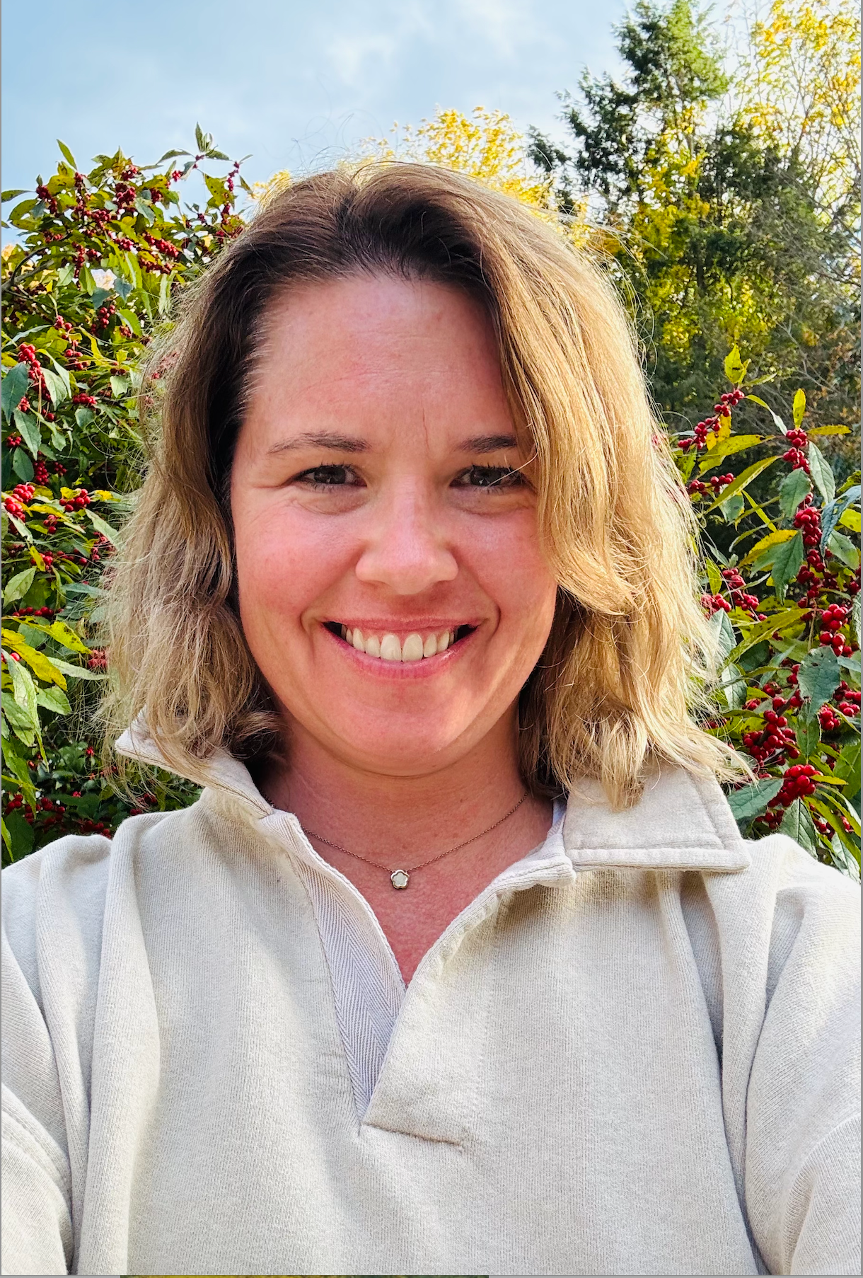 A woman in a white sweater is smiling in front of a bush with red berries.