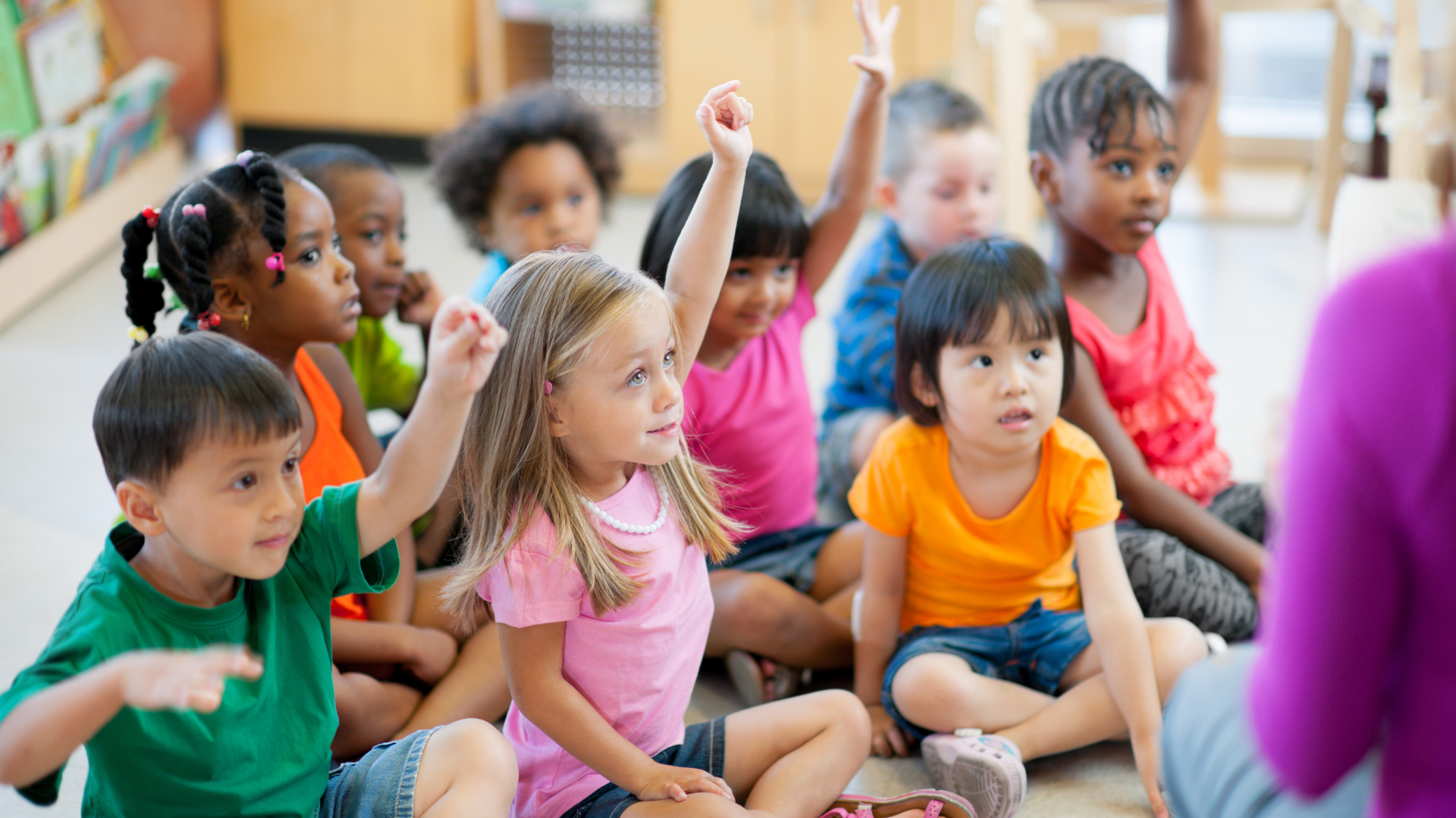A group of children are sitting on the floor raising their hands to answer a question.