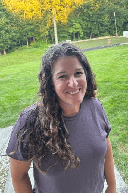 A woman in a purple shirt is standing in front of a lush green field.