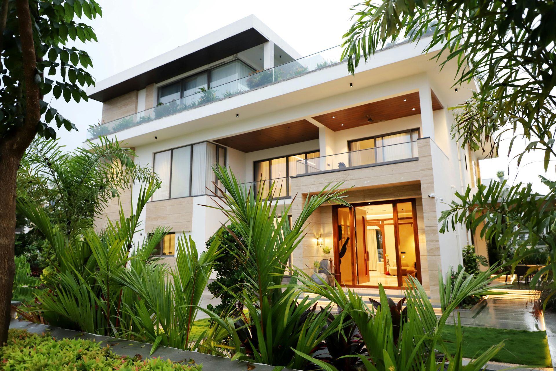 A modern, three-story white villa with glass balconies and floor-to-ceiling windows, surrounded by lush tropical plants.