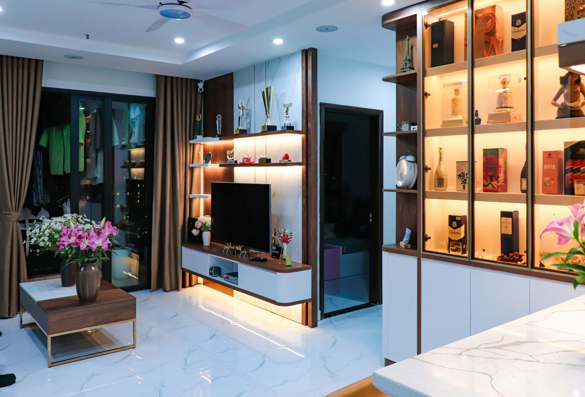 A modern living room with a TV, display shelves featuring bottles, beige curtains, and a low coffee table with flowers.