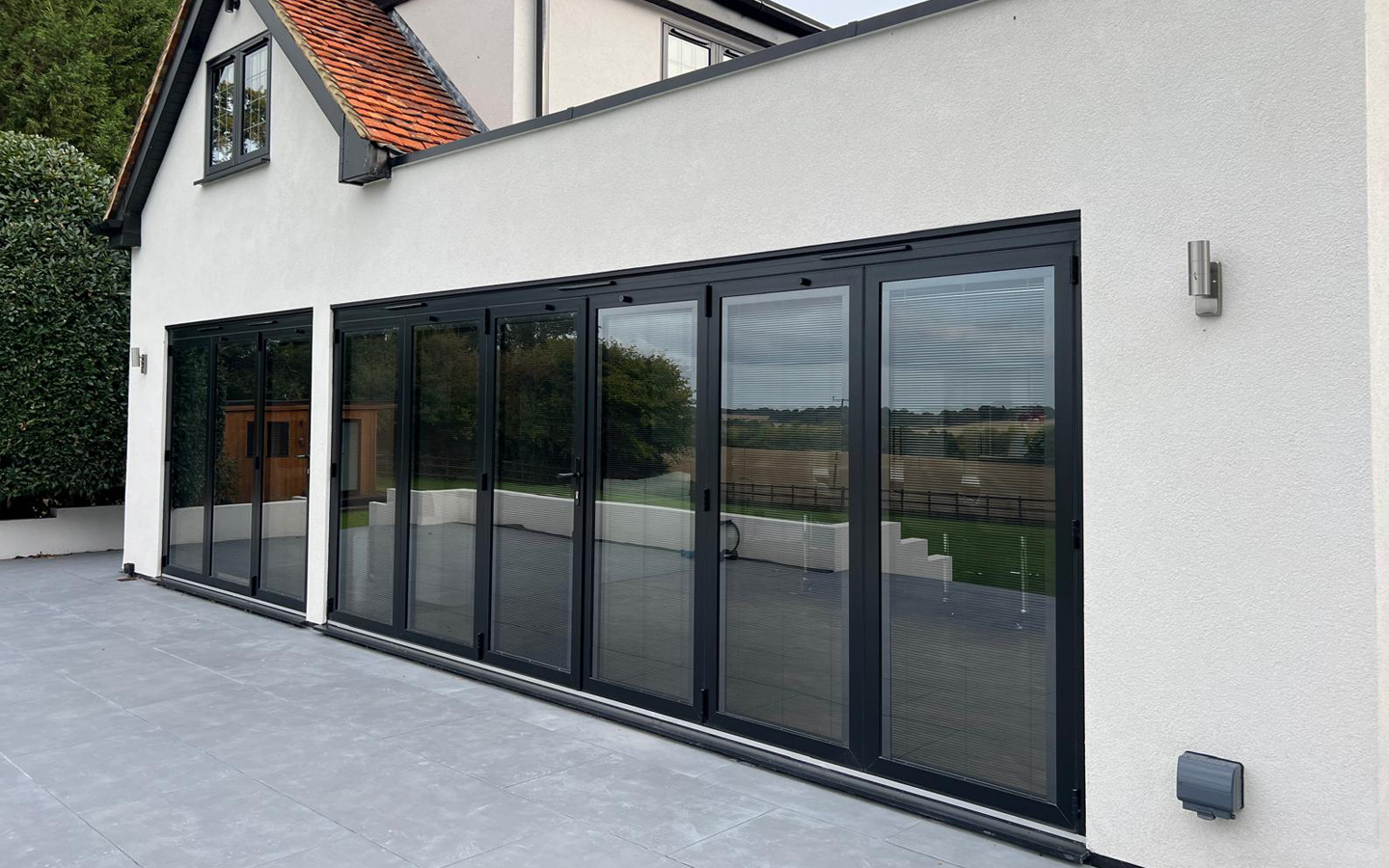 Modern home exterior with large black-framed folding glass doors, white walls, and a gray patio.