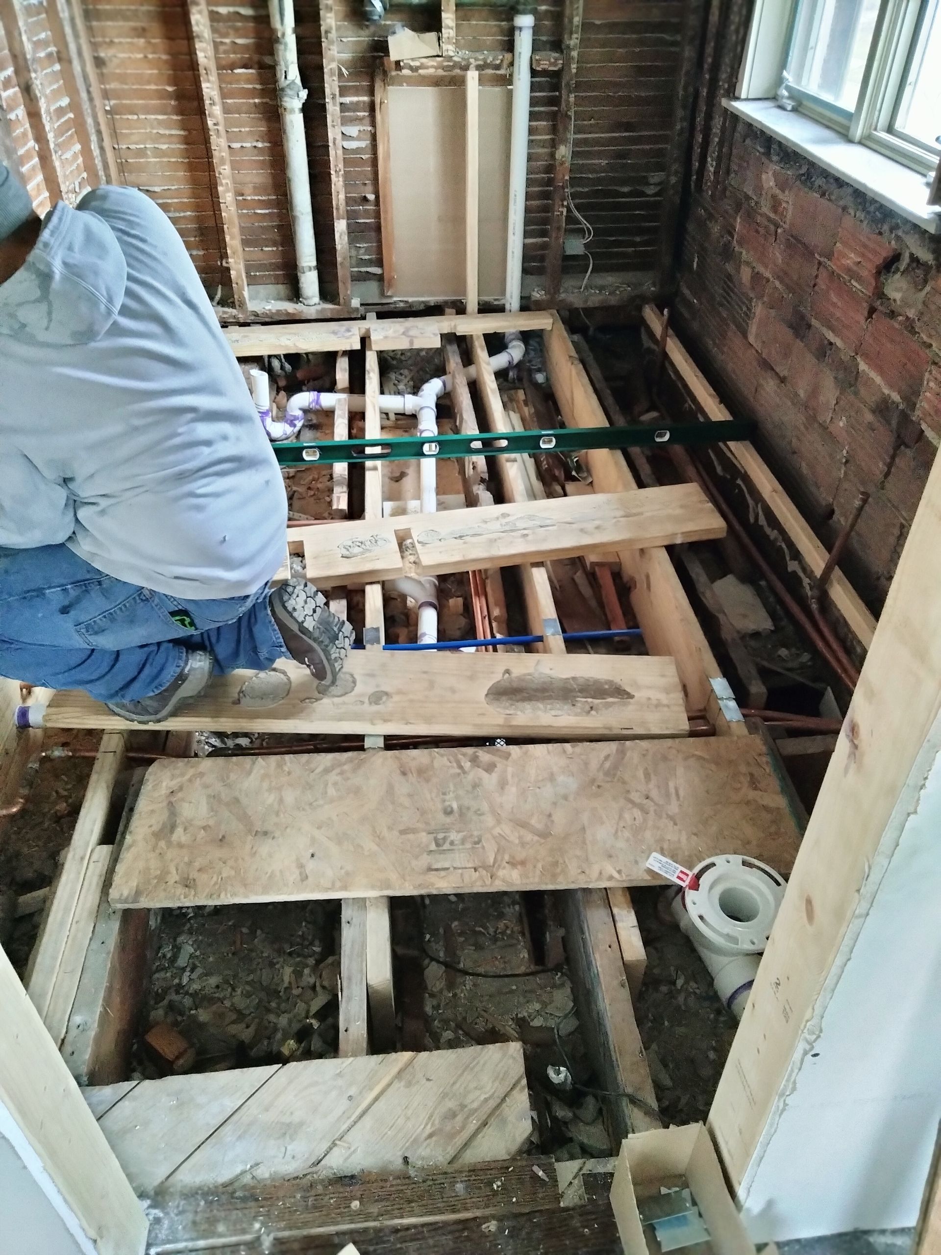 A man is kneeling on a wooden floor in a bathroom under construction.