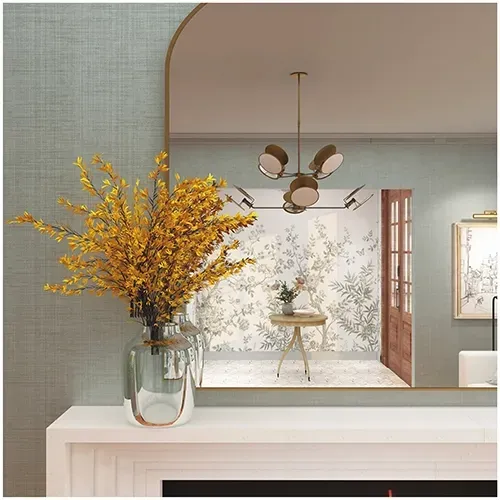 A clear glass vase filled with bright yellow branches sits on a white mantel in front of an arched mirror.