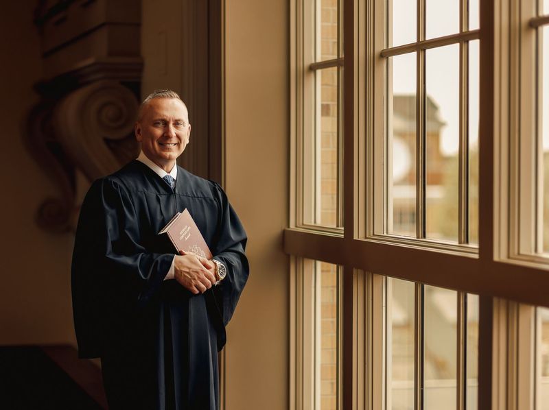 A man in a graduation gown is standing in front of a window holding a book.