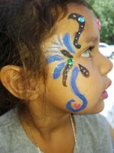 A little girl has her face painted with a dragonfly design.