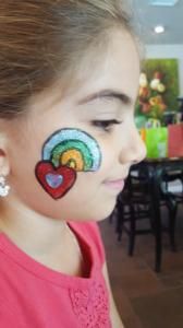 A little girl with a rainbow and two hearts painted on her face.