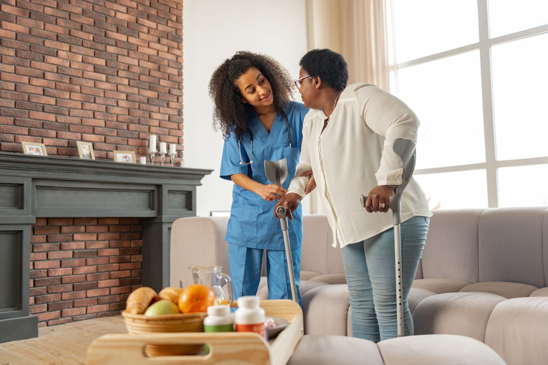 A nurse is helping a woman with crutches walk in a living room.